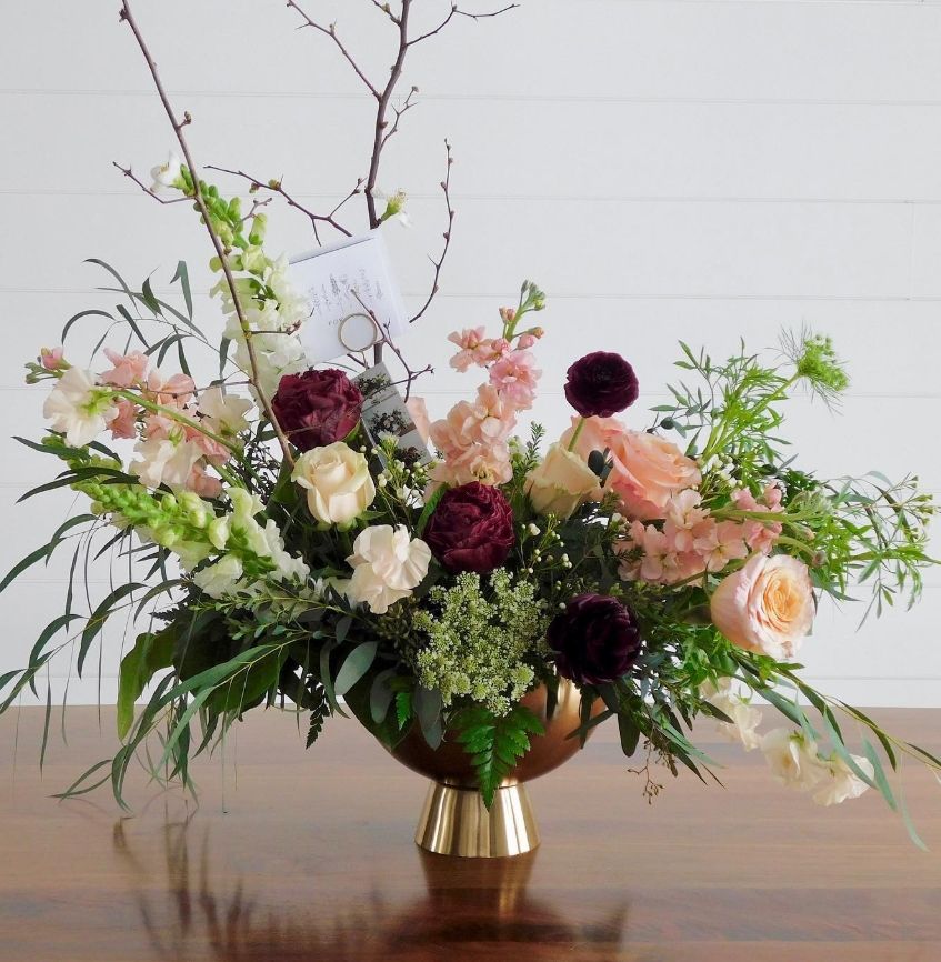 Elegant floral arrangement with pink roses, dark burgundy blooms, and delicate greenery in a gold metallic vase.