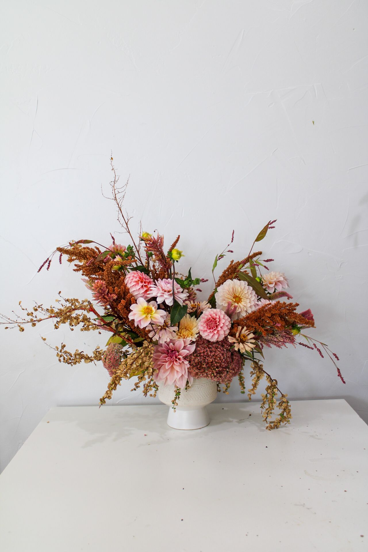 Asymmetrical floral design with dried elements, pink blooms and golden branches in a white container.