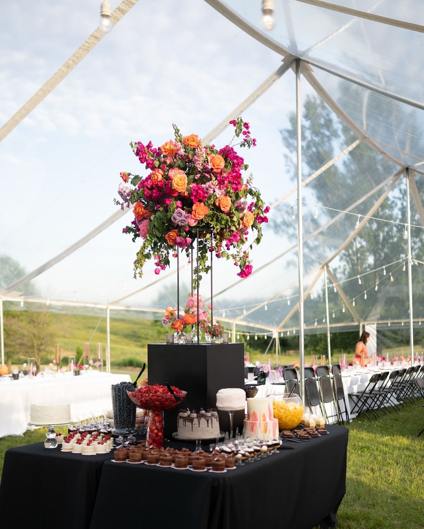 Elegant outdoor wedding reception buffet table with tall floral centerpiece under clear tent canopy.