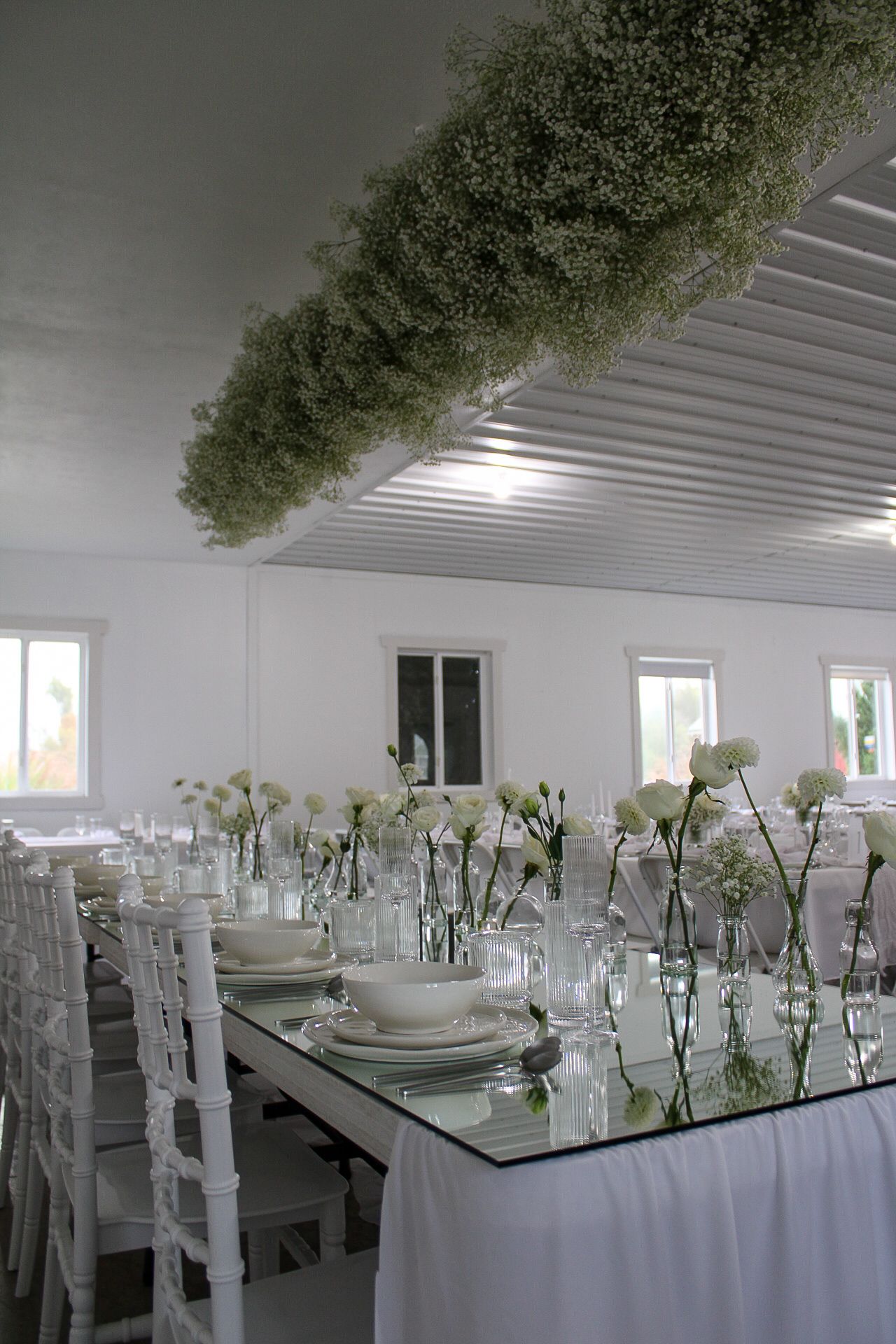 Modern wedding table setup with white chairs and delicate floral arrangements under a greenery installation.