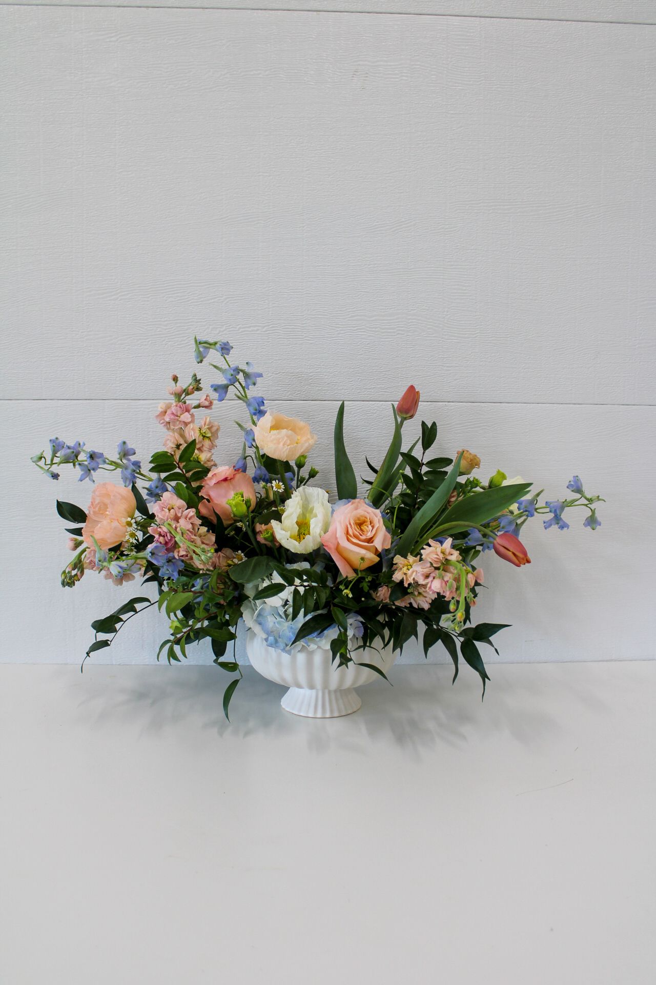 A garden-inspired floral arrangement with peach roses, blue delphinium and tulips in a white pedestal vase.