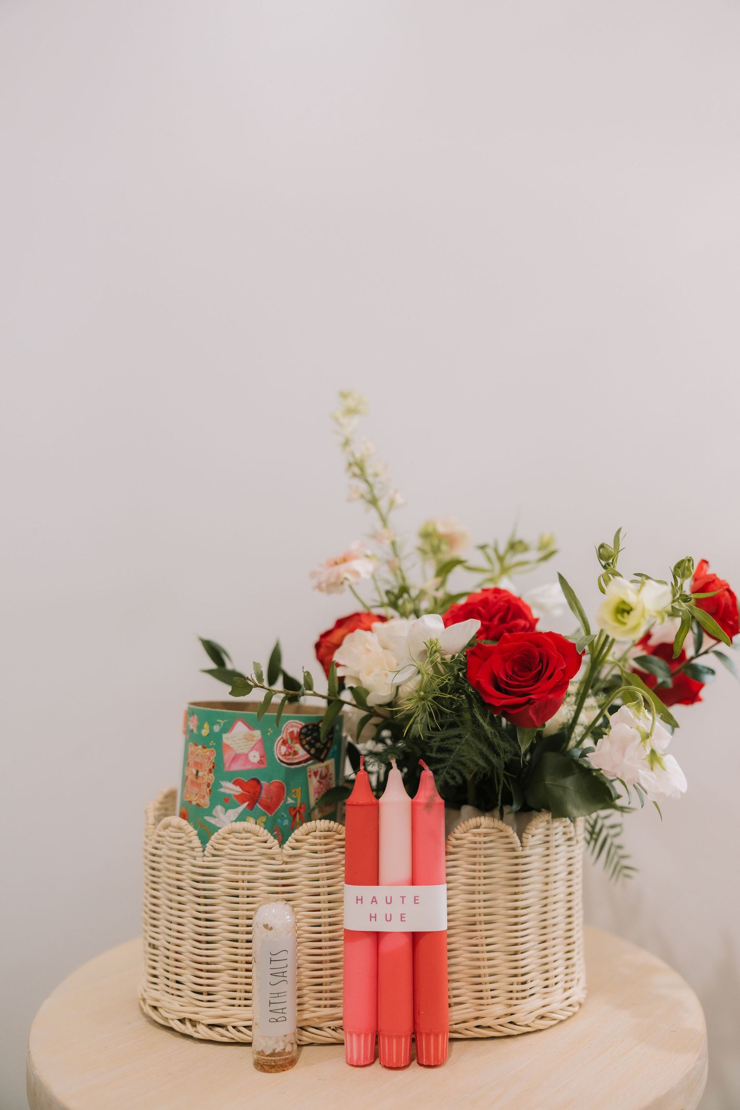 A wicker basket arrangement with red roses, white flowers, pink candles and a decorative green tin on a light surface.