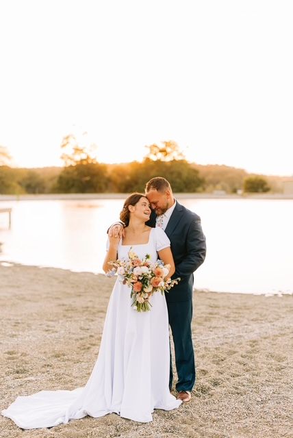 Romantic wedding couple embraces on a sandy beach at sunset with flowing white dress and bouquet.