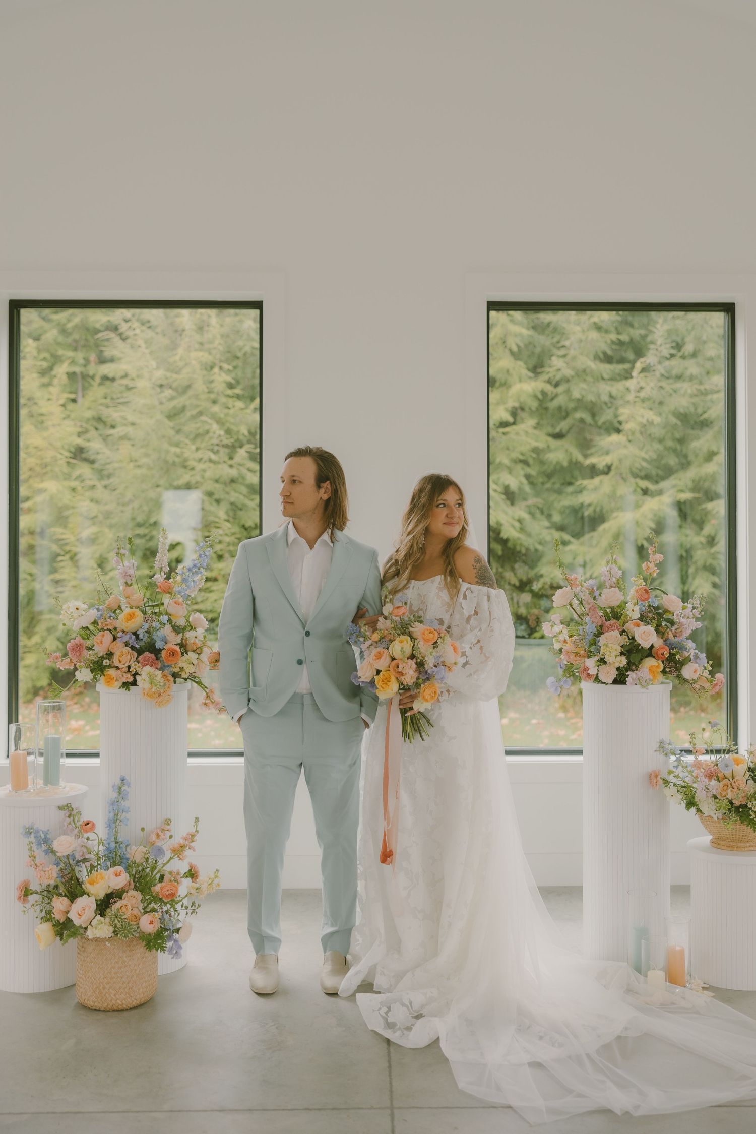 A wedding couple poses in a bright white room surrounded by floral arrangements and pedestals.