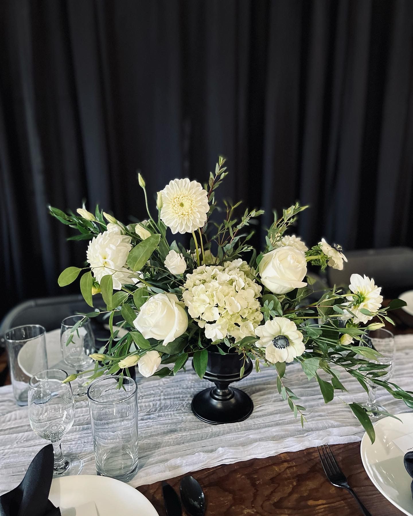 White floral centerpiece with hydrangeas and roses in black pedestal vase on marble-style table.