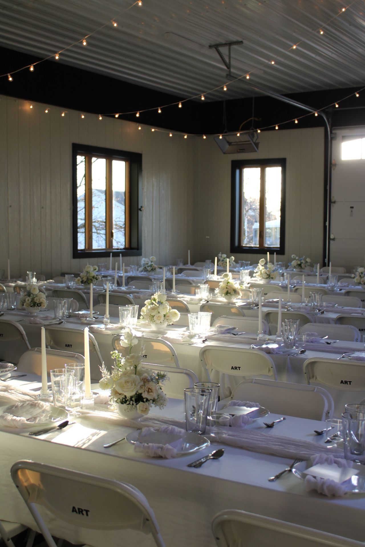 Wedding reception venue with string lights and white table settings arranged in long dining rows.