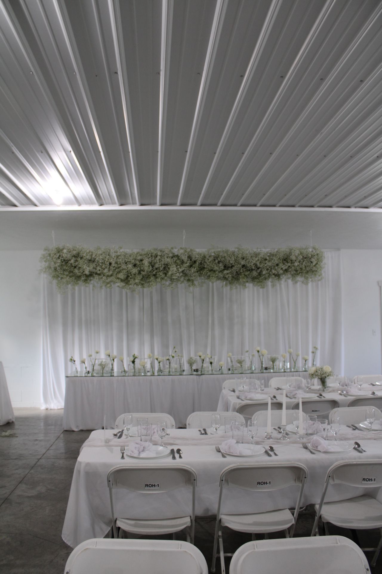 Wedding reception space with white folding chairs and a dramatic greenery installation overhead.