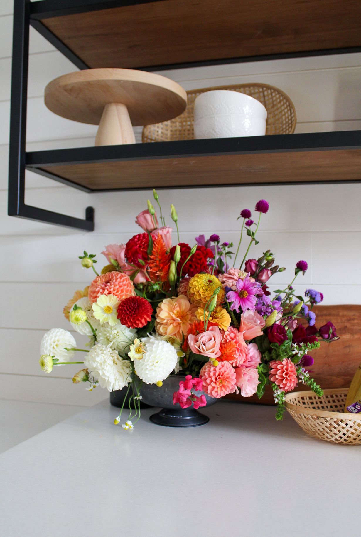 Colorful floral arrangement with dahlias and roses on shelf with decorative mushroom sculptures.