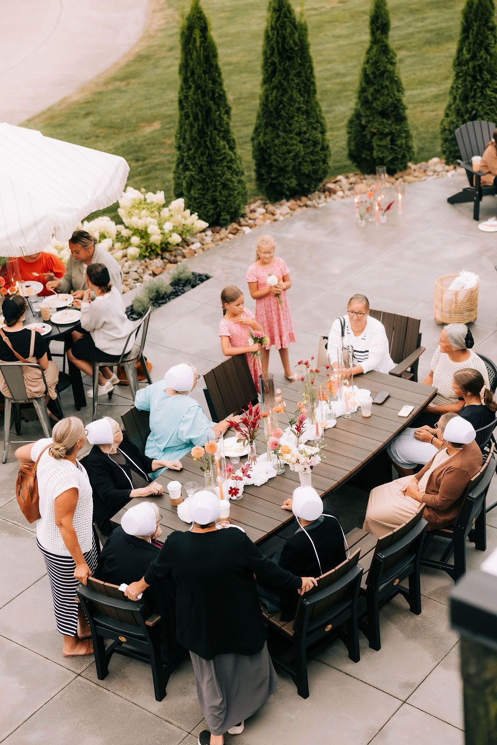 Guests seated at long outdoor dining tables on a patio surrounded by cypress trees during a social gathering.