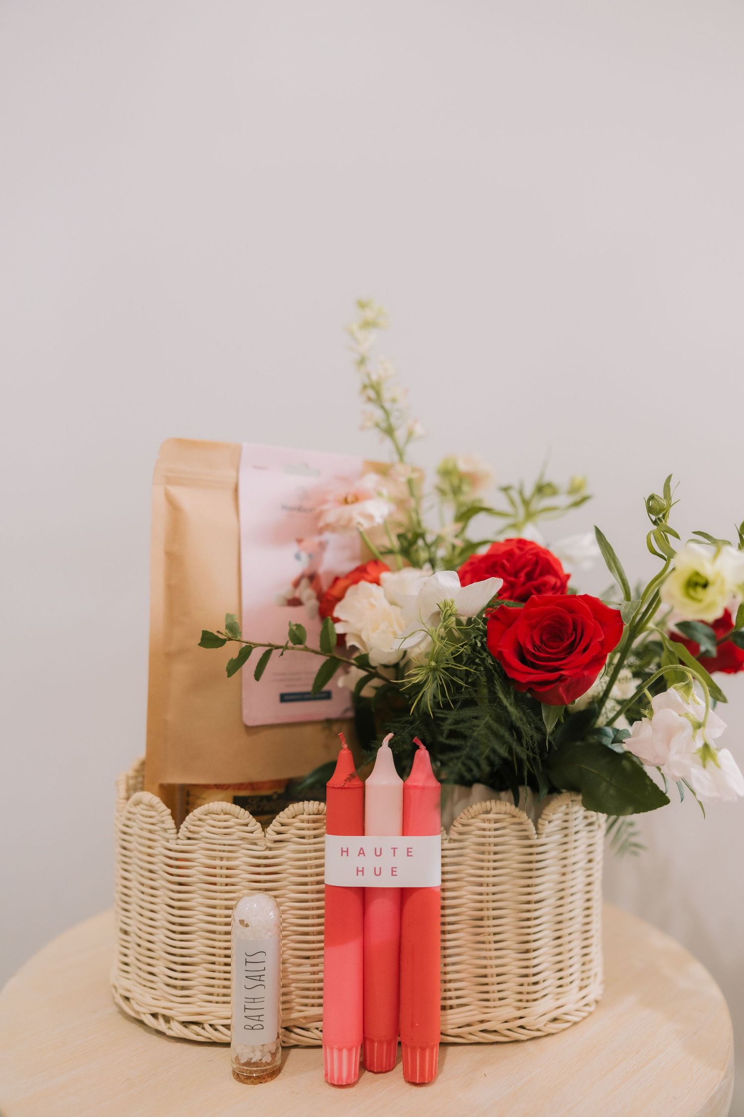 Wicker basket filled with red roses and white flowers alongside pink candles and decorative items on a light surface.