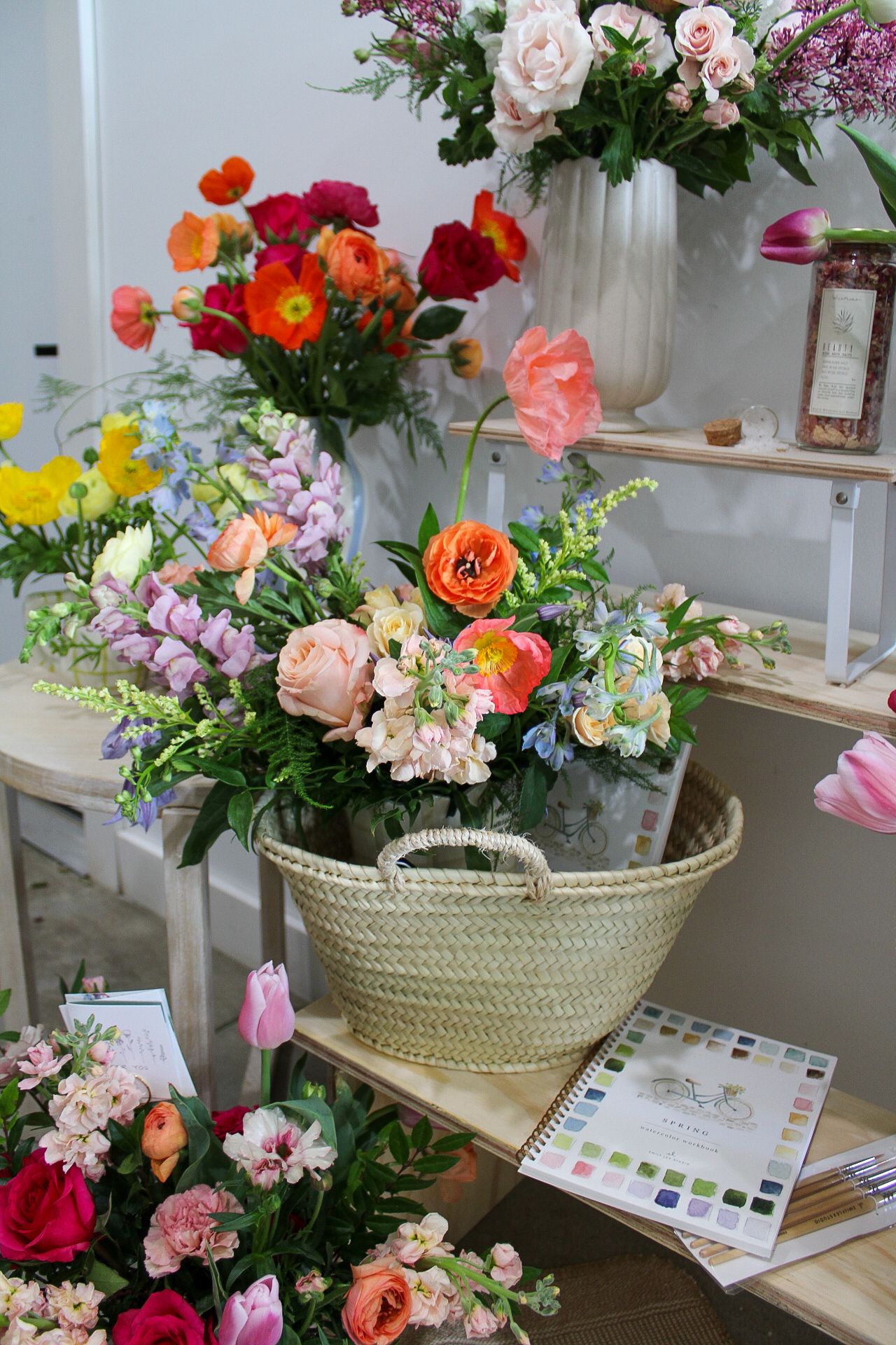 Colorful floral arrangements with roses, poppies, and wildflowers displayed on wooden shelves with a woven basket centerpiece.