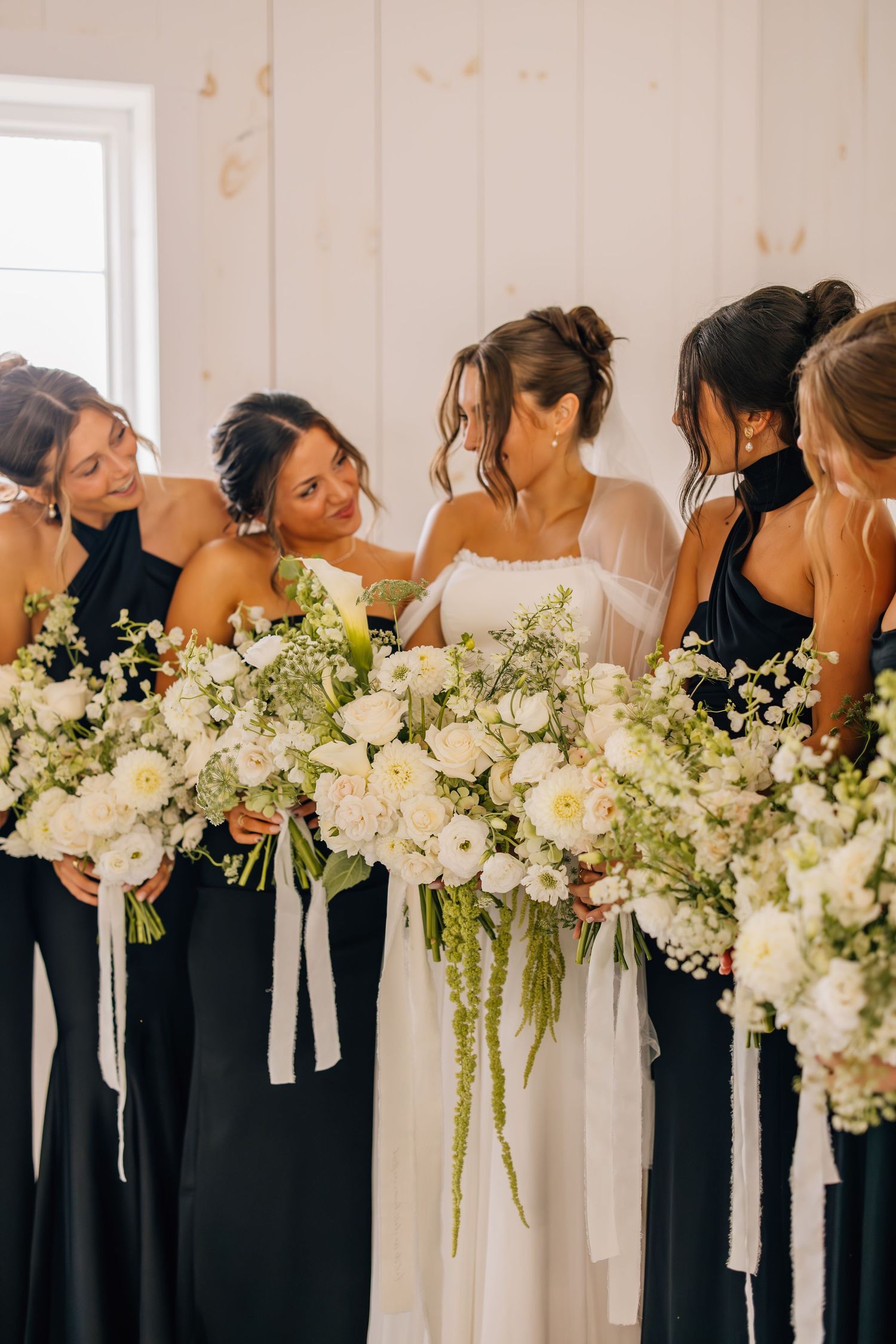 A row of bridesmaids in black dresses hold elegant white and green floral bouquets against a white wall.