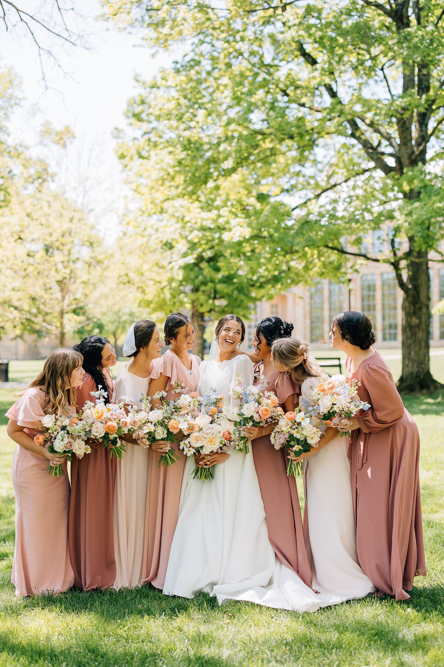 Bridesmaids in blush and mauve dresses stand in a line holding floral bouquets on a sunny lawn.
