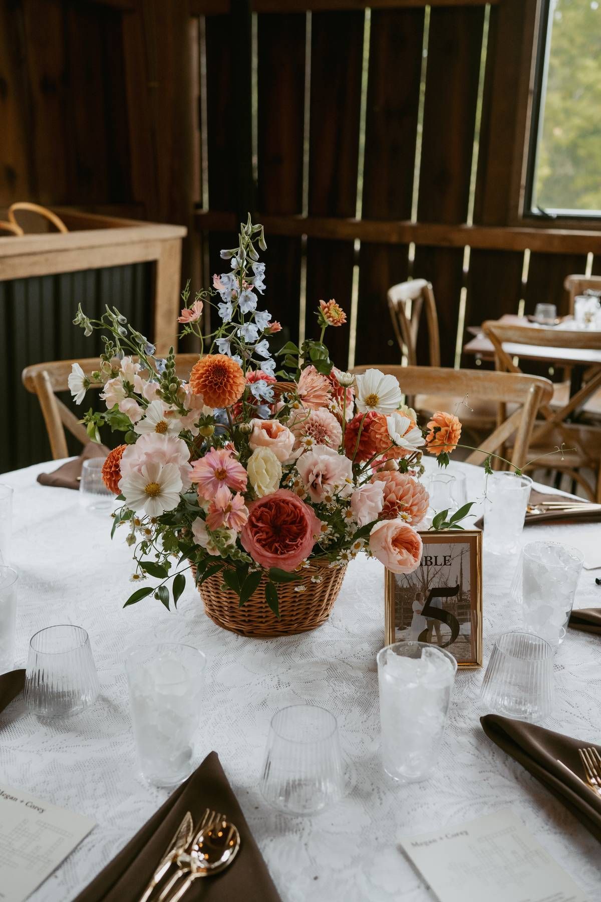 Elegant wedding reception table with coral and pink floral centerpieces on white tablecloth in rustic wooden venue.