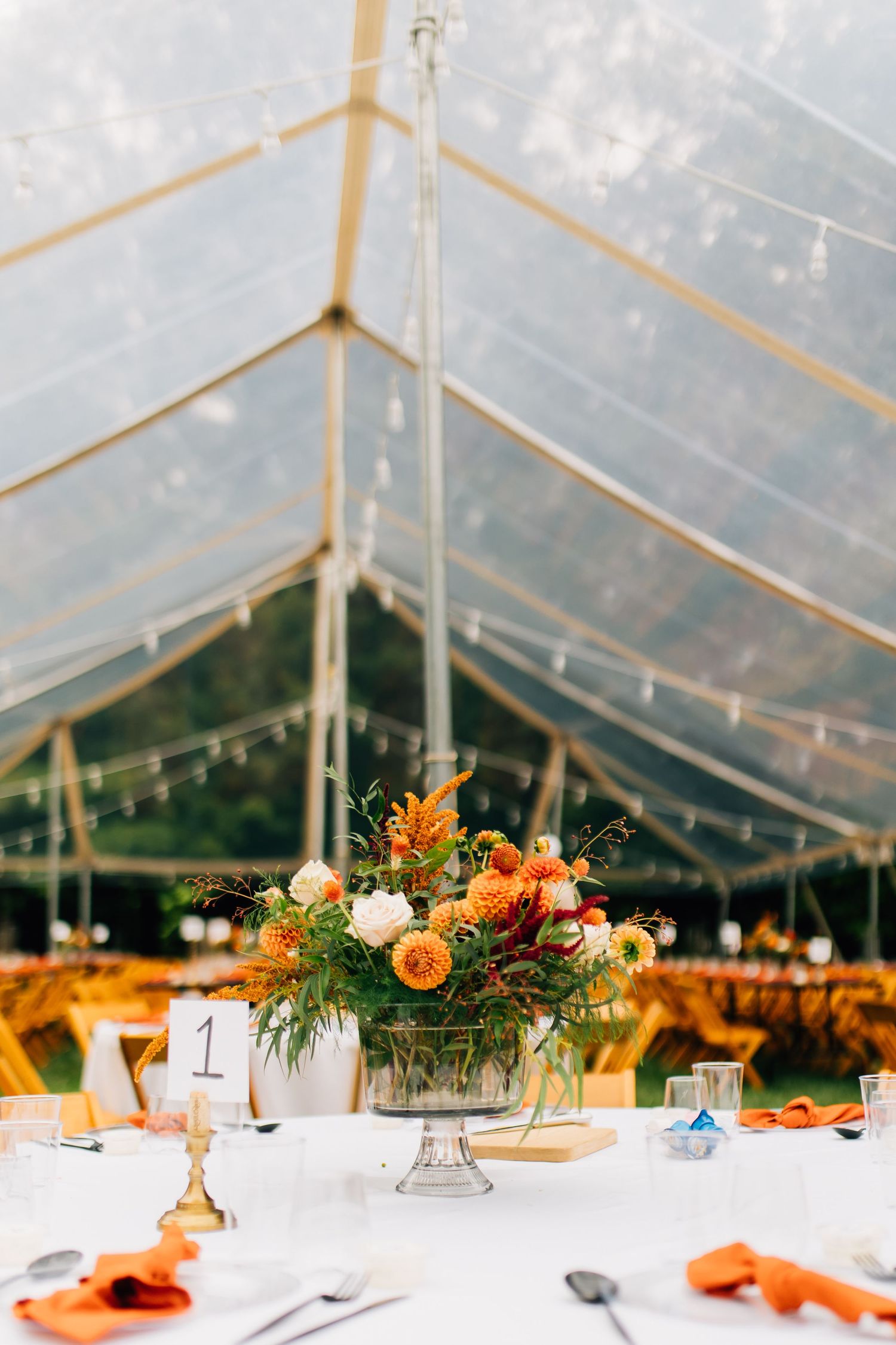 Clear tent wedding reception with orange floral centerpiece and place settings.