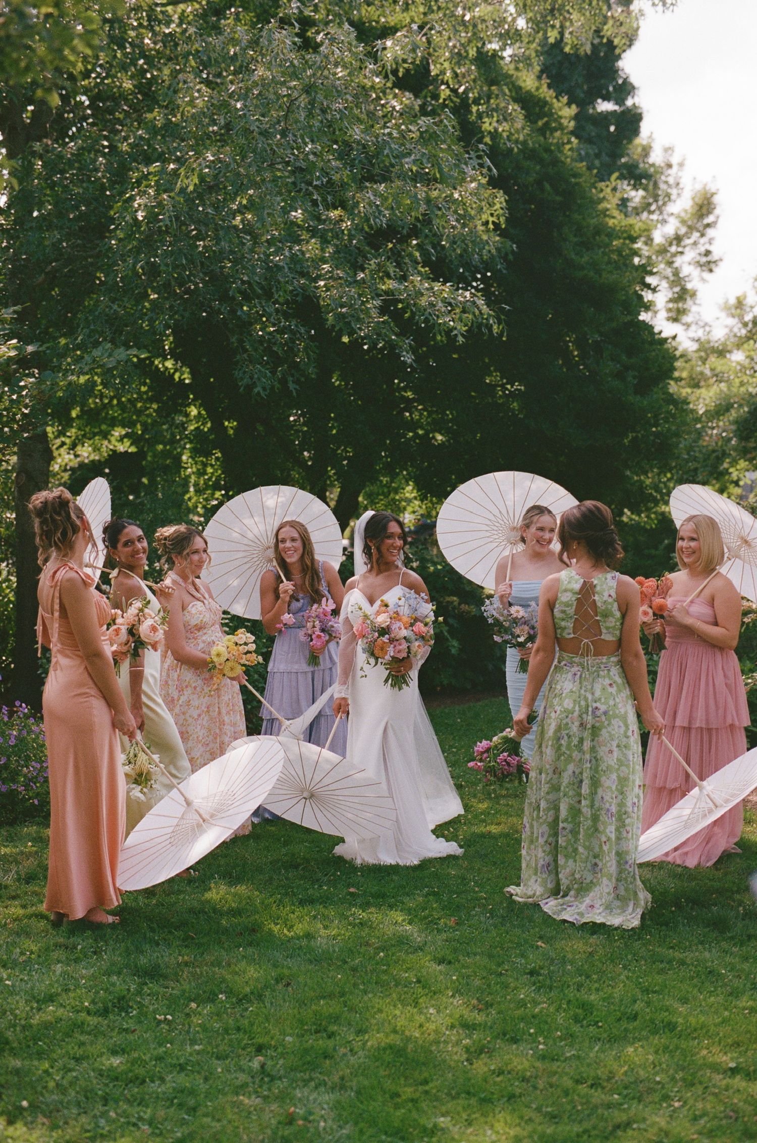 Wedding party with bridesmaids in pastel dresses holding white parasols on a sunny lawn.