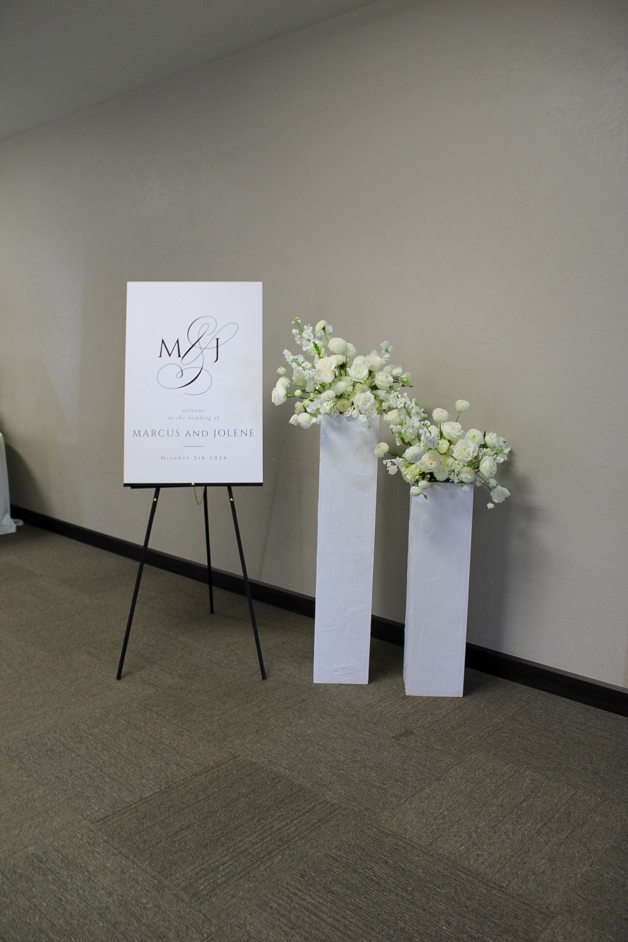 Minimalist wedding signage display with white pedestals holding simple floral arrangements.