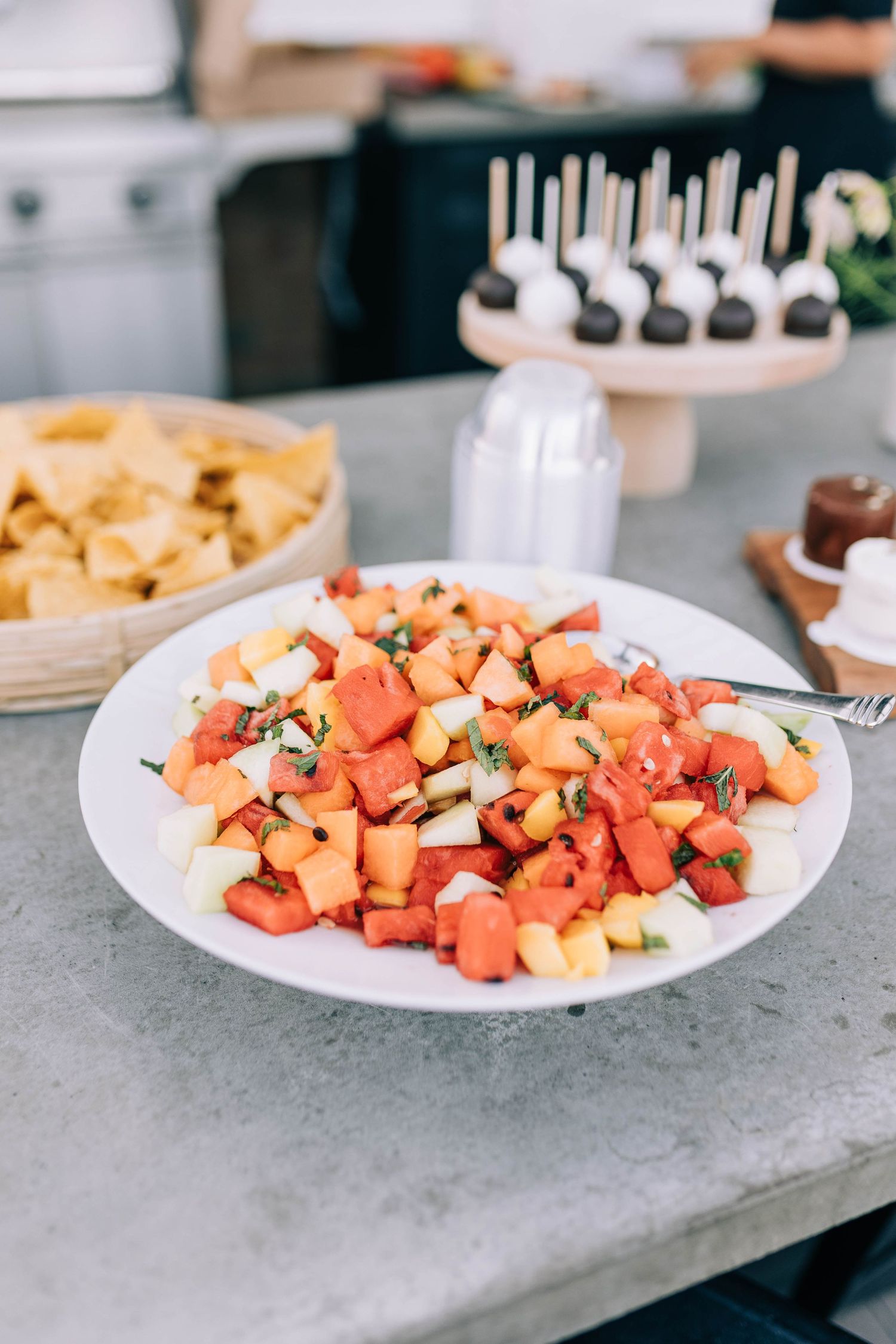 Fresh fruit salad with watermelon, cantaloupe and herbs served on white plate with desserts in background.