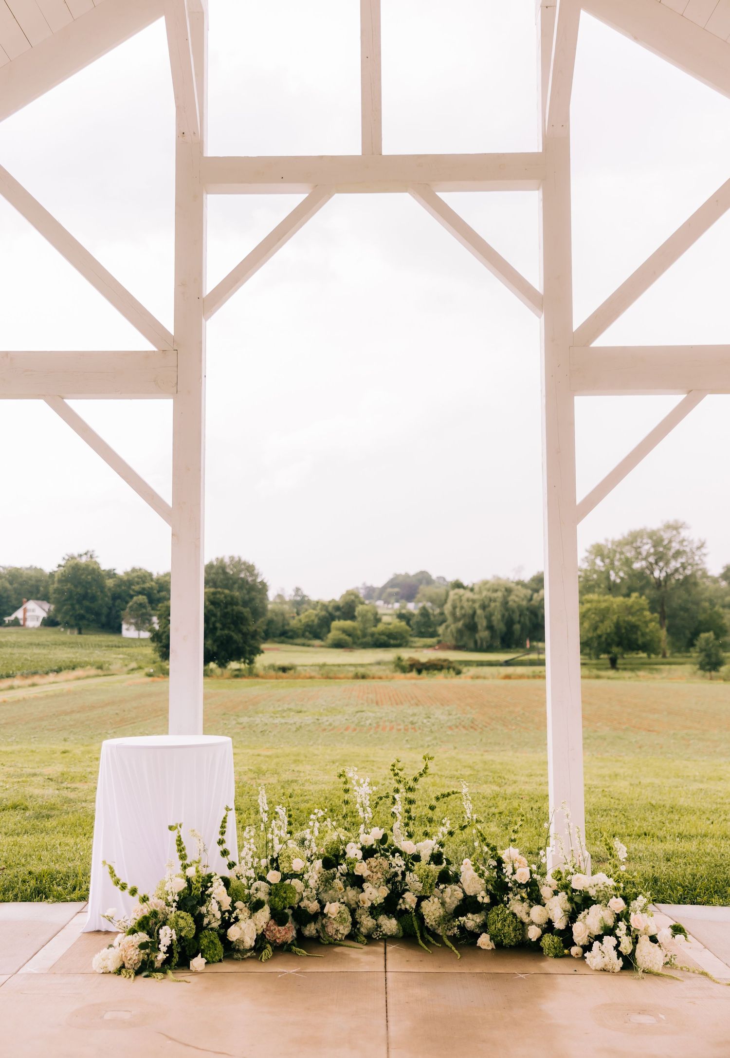 White floral arrangement adorns the base of a modern outdoor wedding pavilion overlooking rolling green countryside.