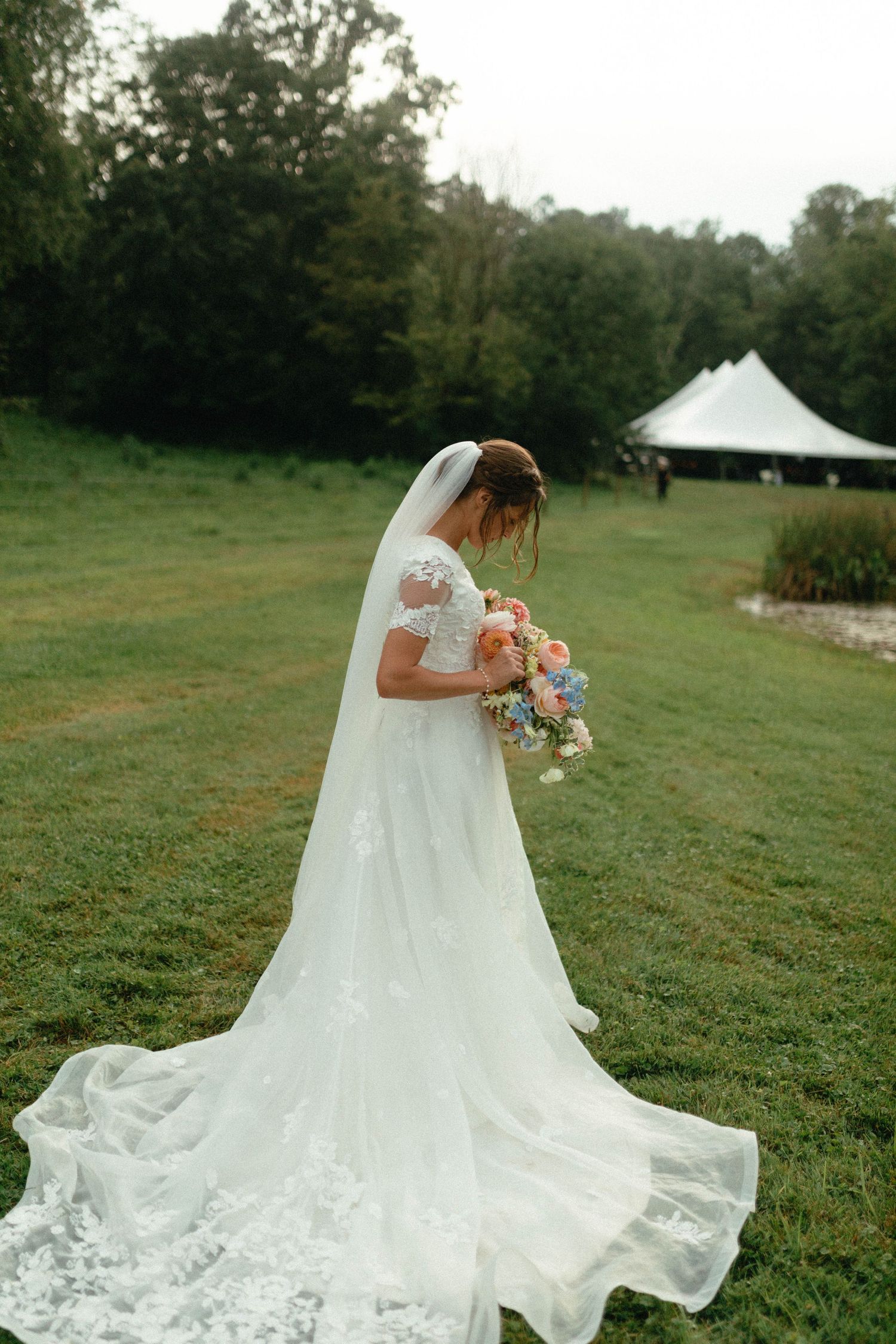 A bride in a flowing white lace wedding dress stands in a green field with a bouquet of pastel flowers.
