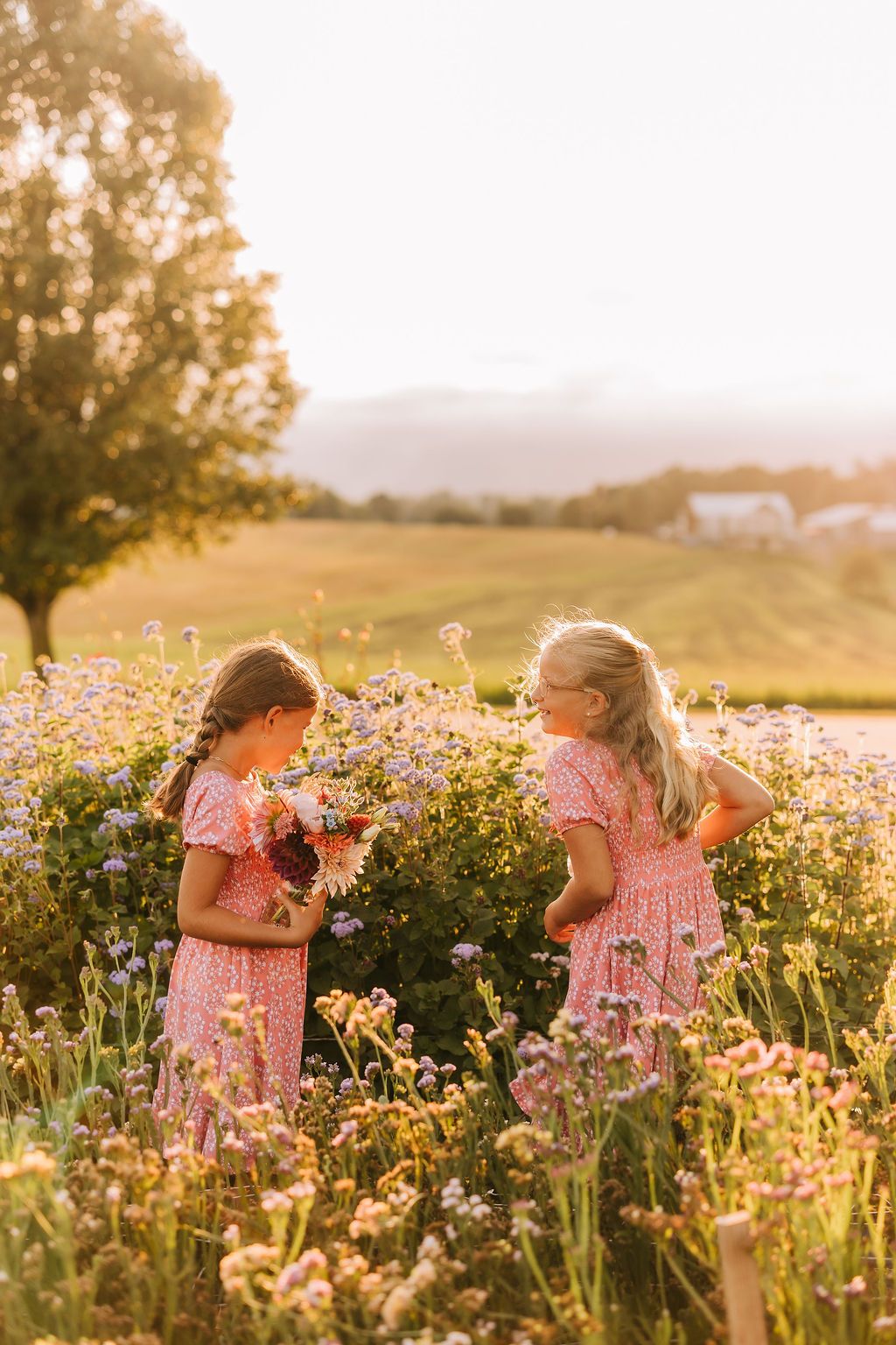 Two children in pink dresses explore a wildflower meadow at golden hour as sunlight streams through the flowers.