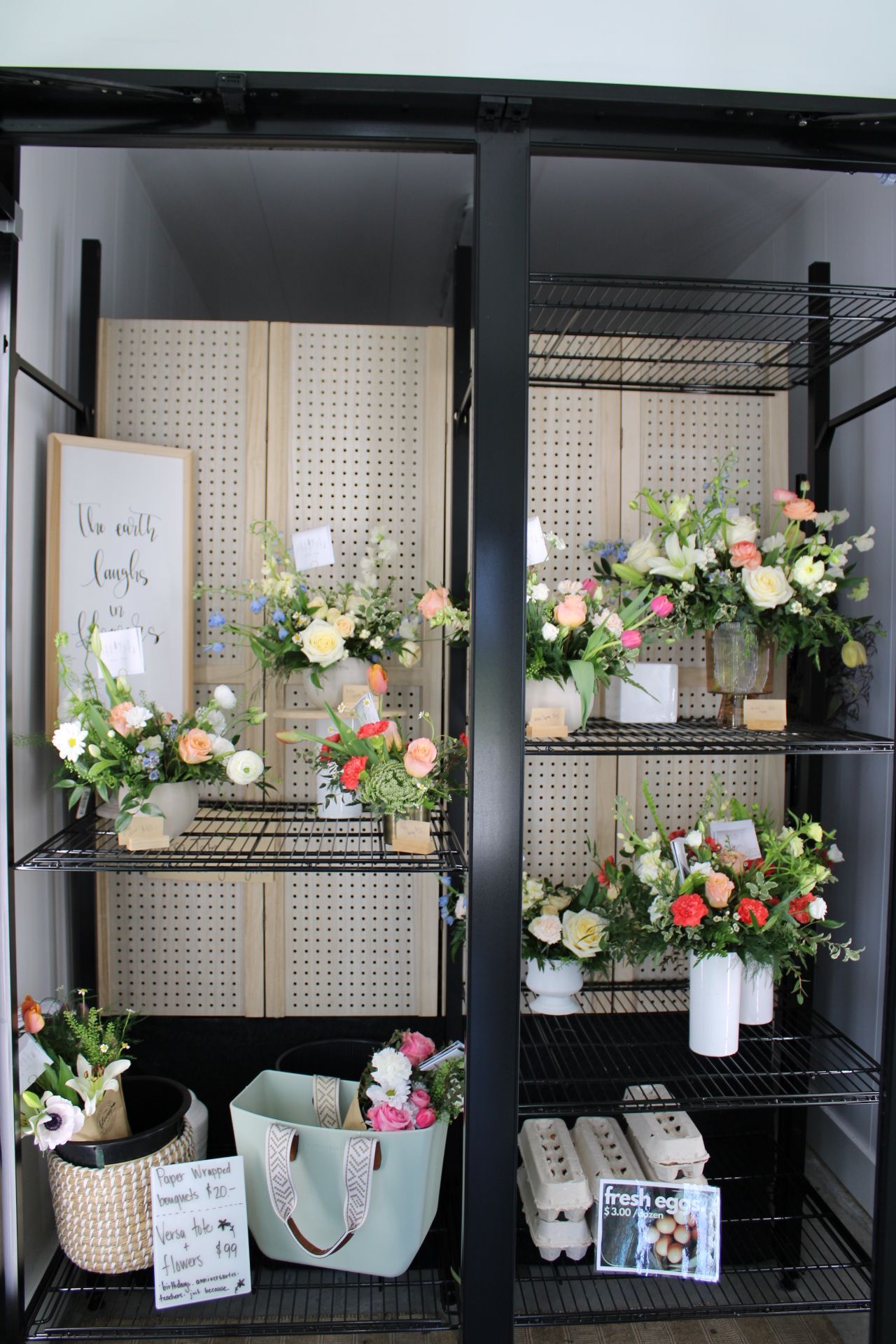 Black retail display unit showcasing fresh flower arrangements and decorative signs on pegboard backing.