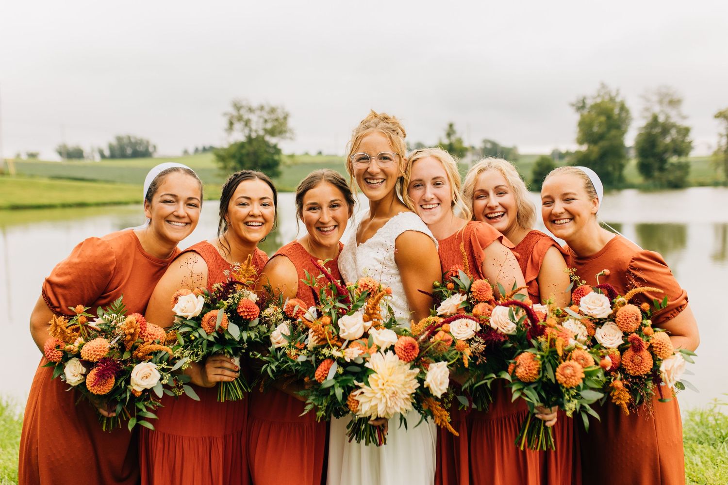 Wedding party in rust-colored dresses poses together by a lake with orange and white bouquets.