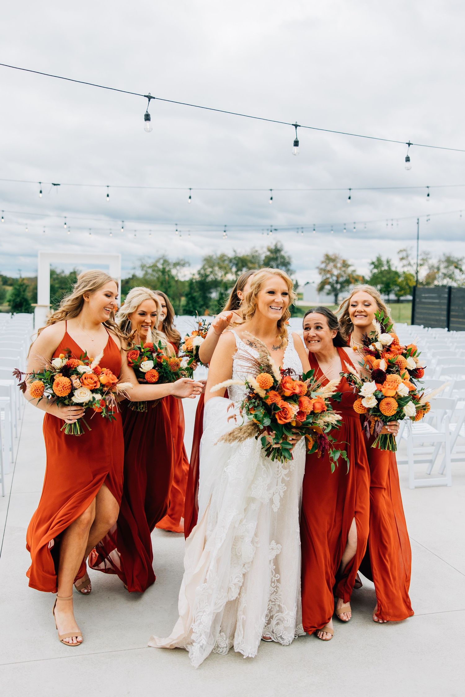 Wedding party in rust colored dresses laughing together while holding vibrant orange and white floral bouquets.