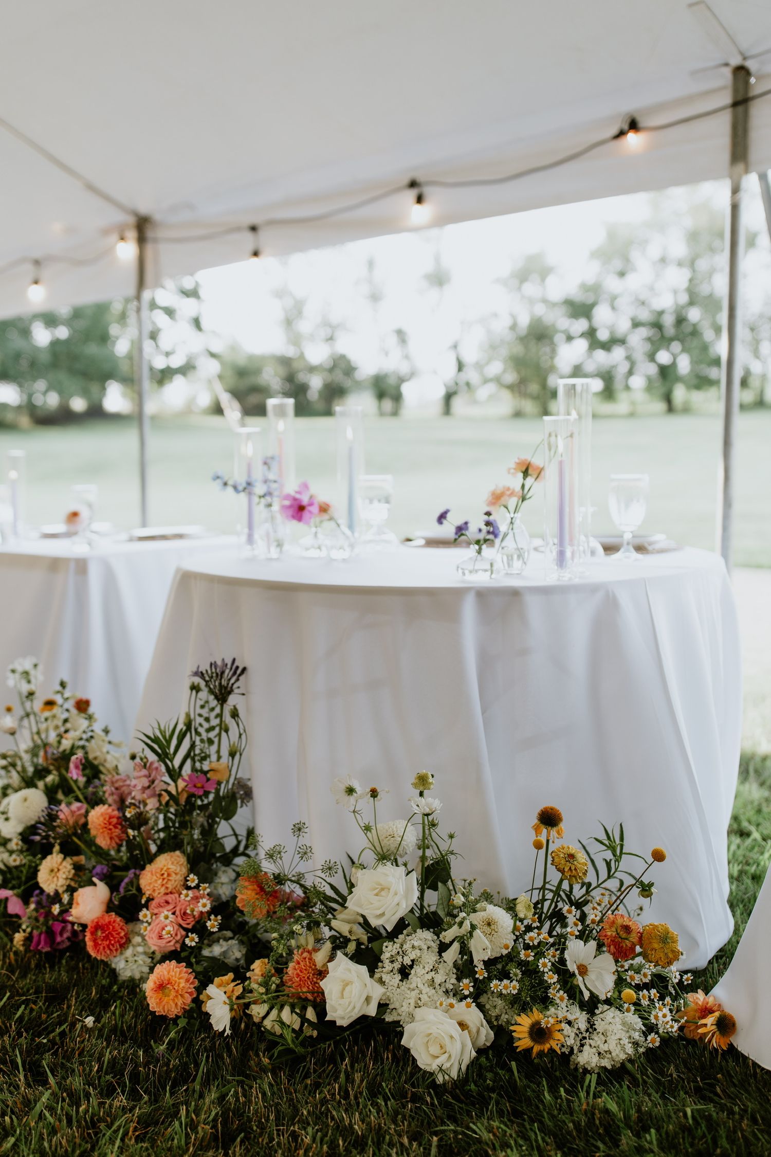 Outdoor wedding reception table with lush floral arrangements under a white tent.