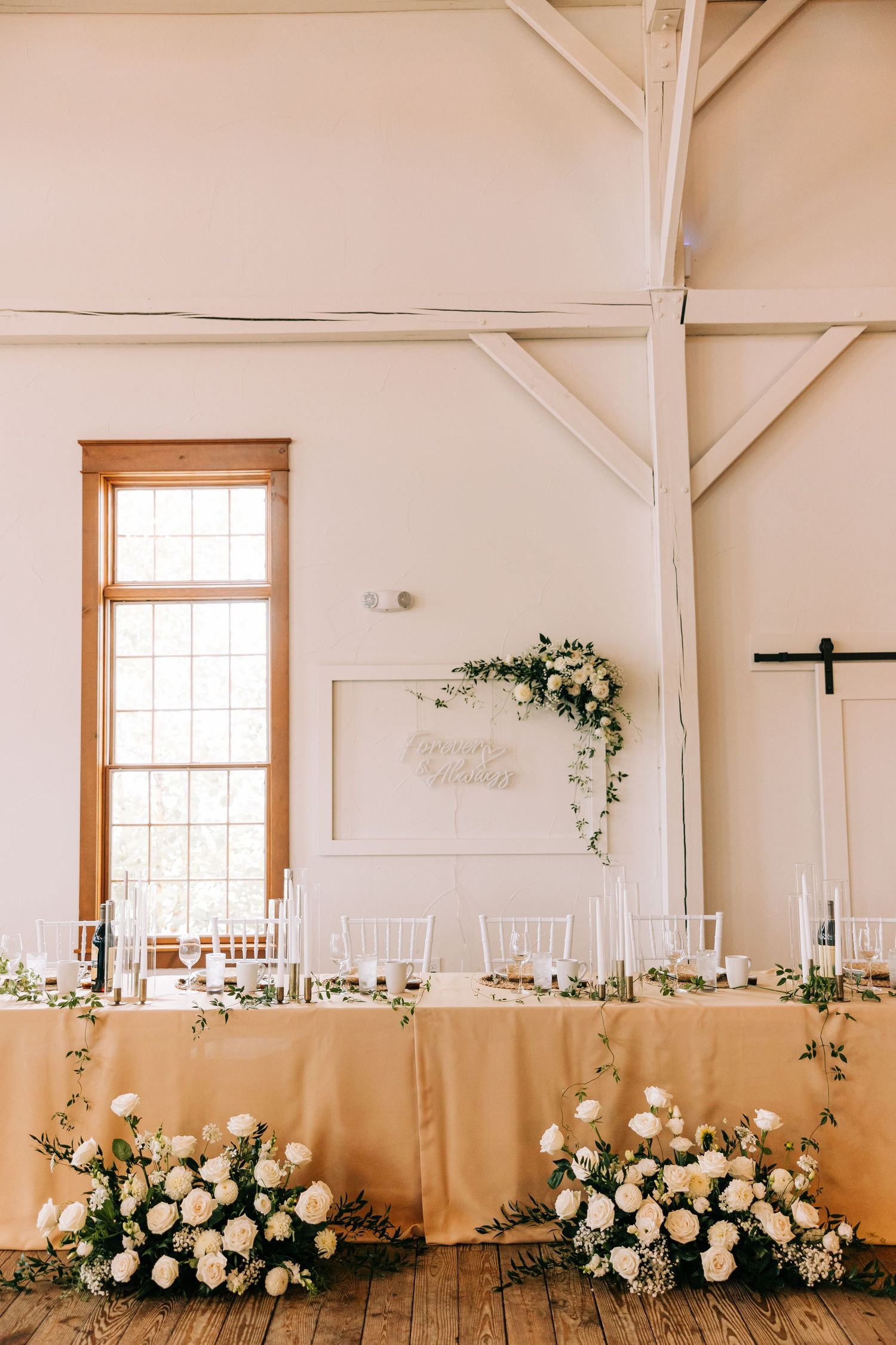 Elegant wedding reception head table with cream tablecloth and white floral arrangements in a bright rustic venue.