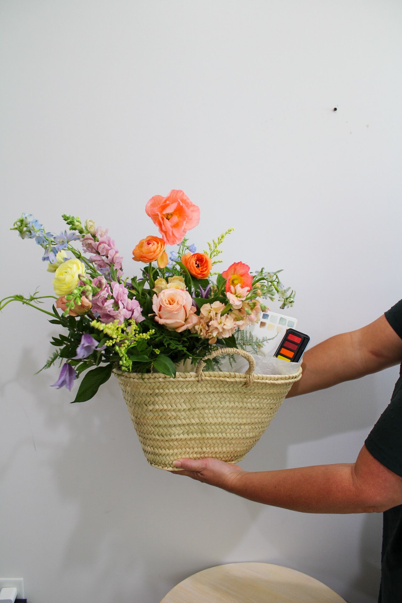 Hand holding a woven straw basket filled with colorful spring flowers including orange ranunculus and pink roses.