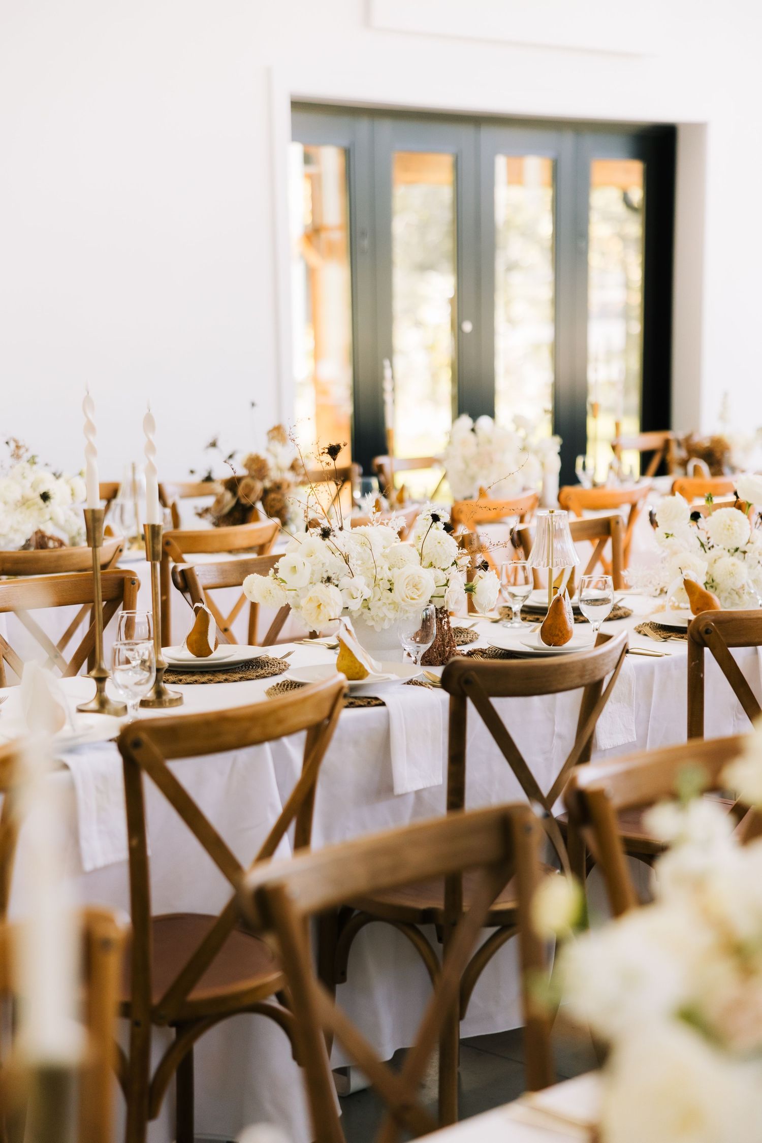 Elegant wedding reception tables set with white linens, wooden cross-back chairs, and delicate floral centerpieces.