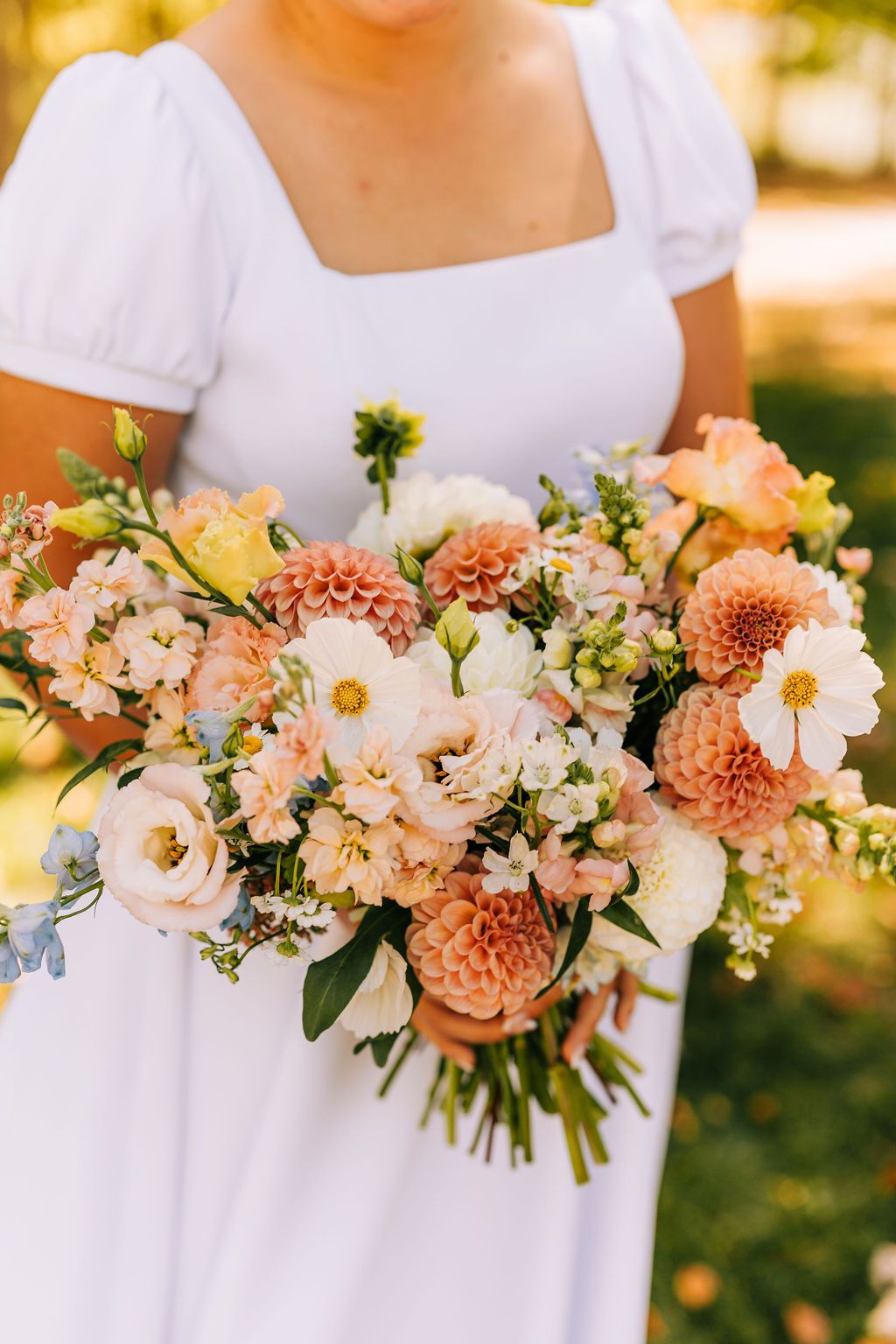 Bridesmaids in white dresses hold elegant peach and coral bouquets with dahlias and mixed flowers in outdoor garden setting.