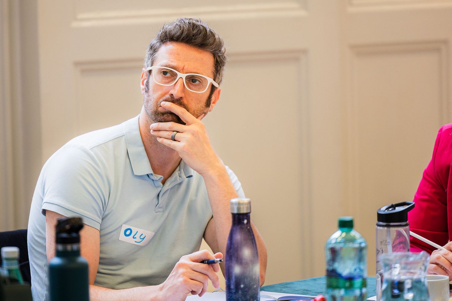Thoughtful man with glasses and beard sitting at a conference table, wearing a light blue polo shirt.