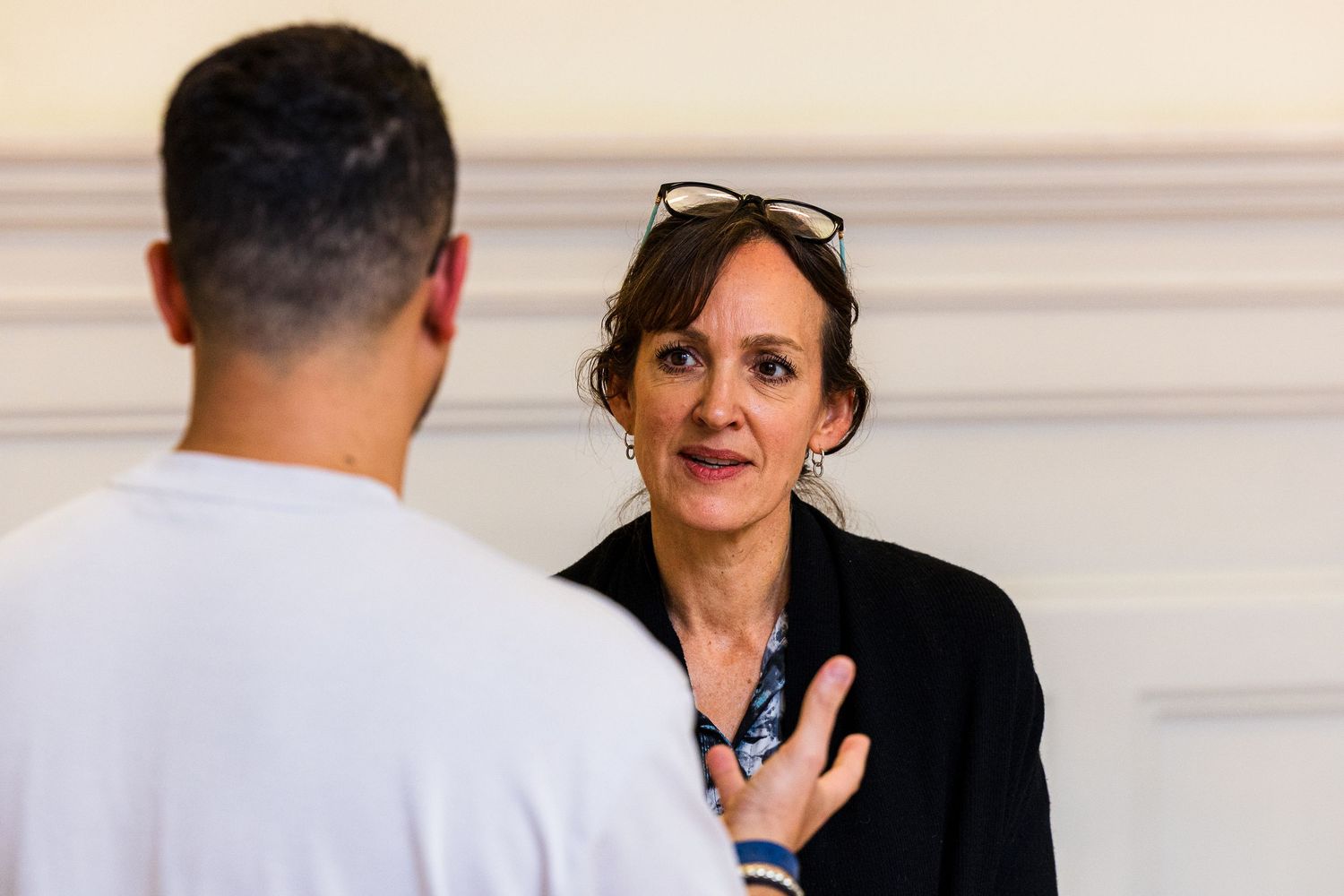 Two people engaged in conversation, woman with sunglasses on head wearing black blazer listening attentively indoors.