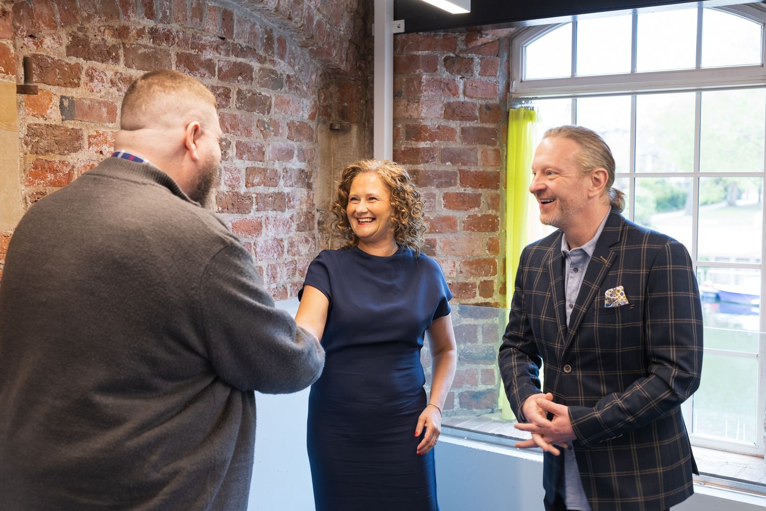 Three professionals networking and smiling in a modern brick-walled office space during a business meeting.