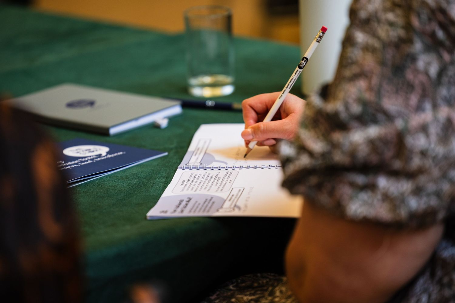 Person writing on a document with a pen at a green table with notebooks and a glass nearby.