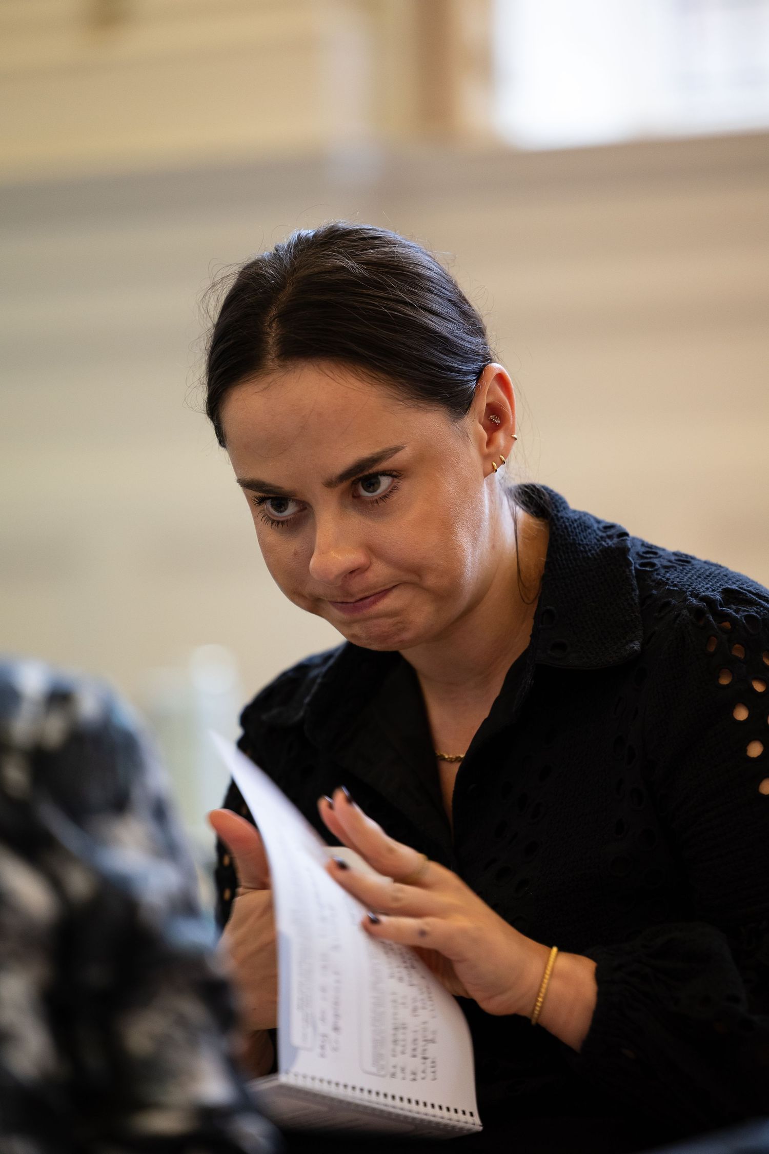 Woman in black shirt holds notepad, looking up with serious expression in bright indoor setting.