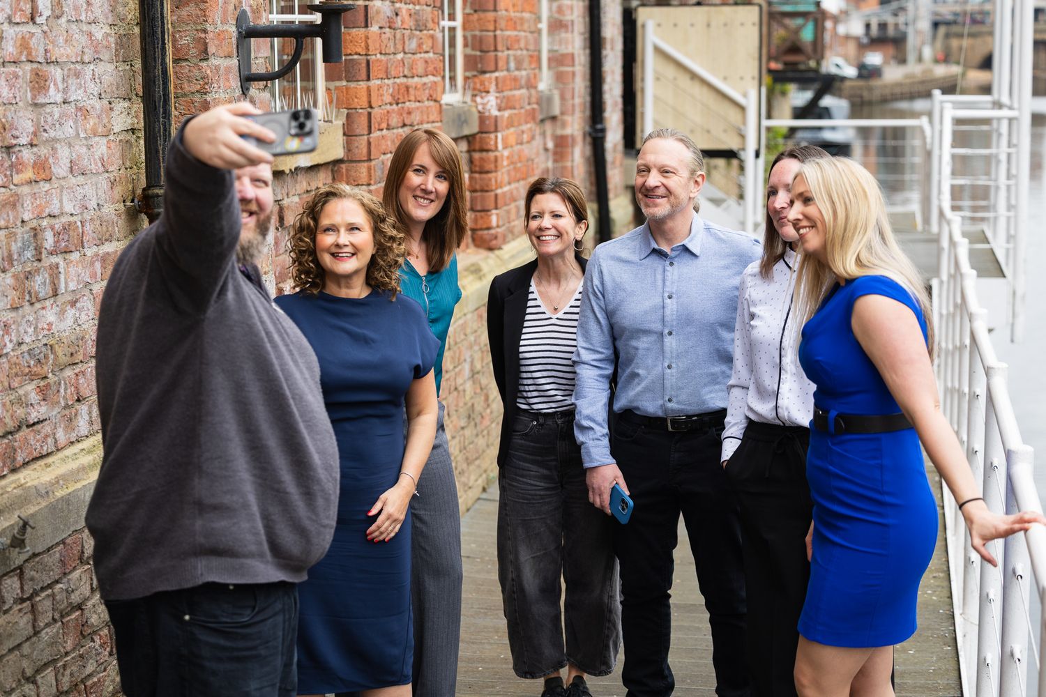 Group of six professionals taking a selfie outdoors against a brick wall building on a sunny day.