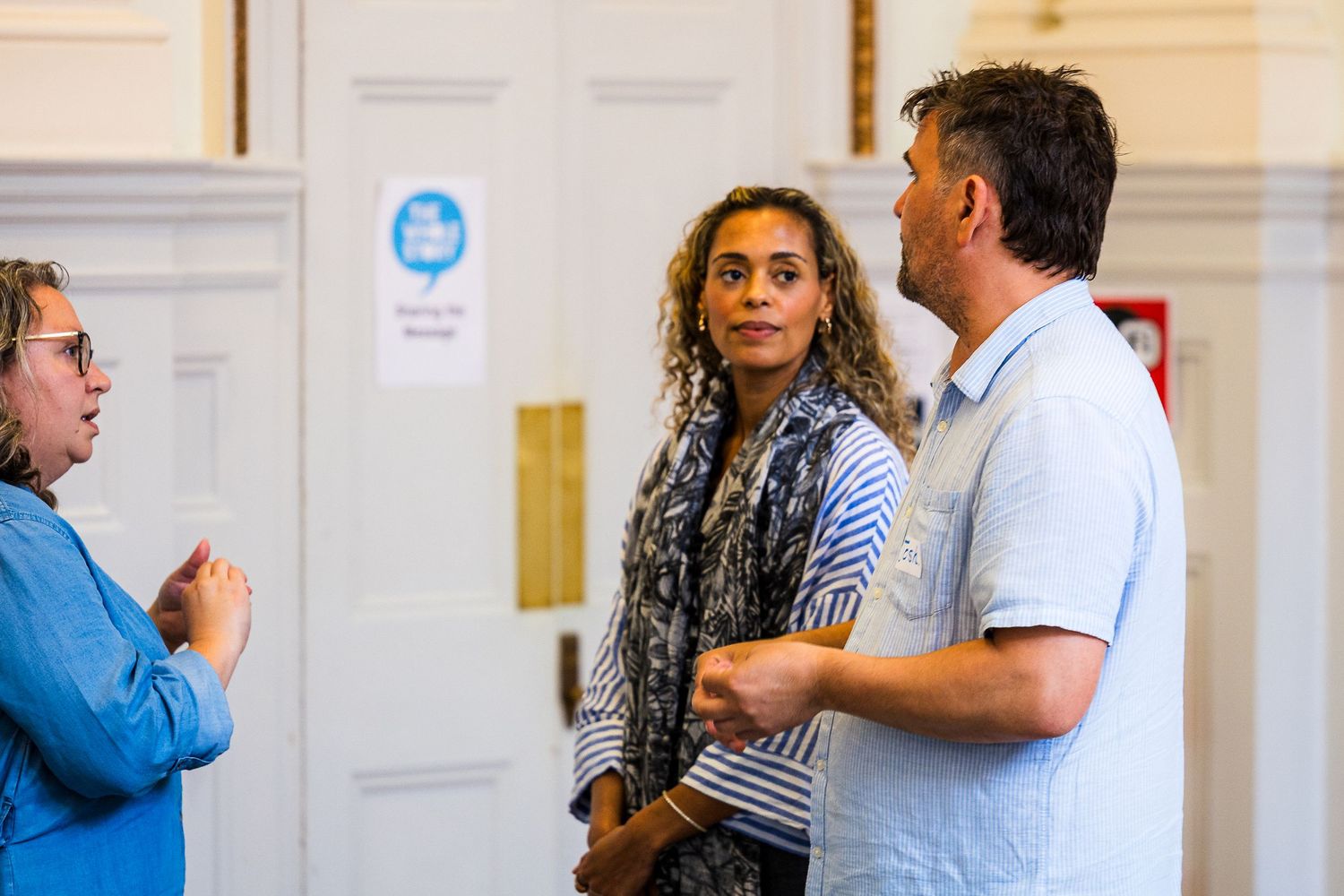 Three people engaged in conversation indoors, two women and a man in casual attire near white doors.