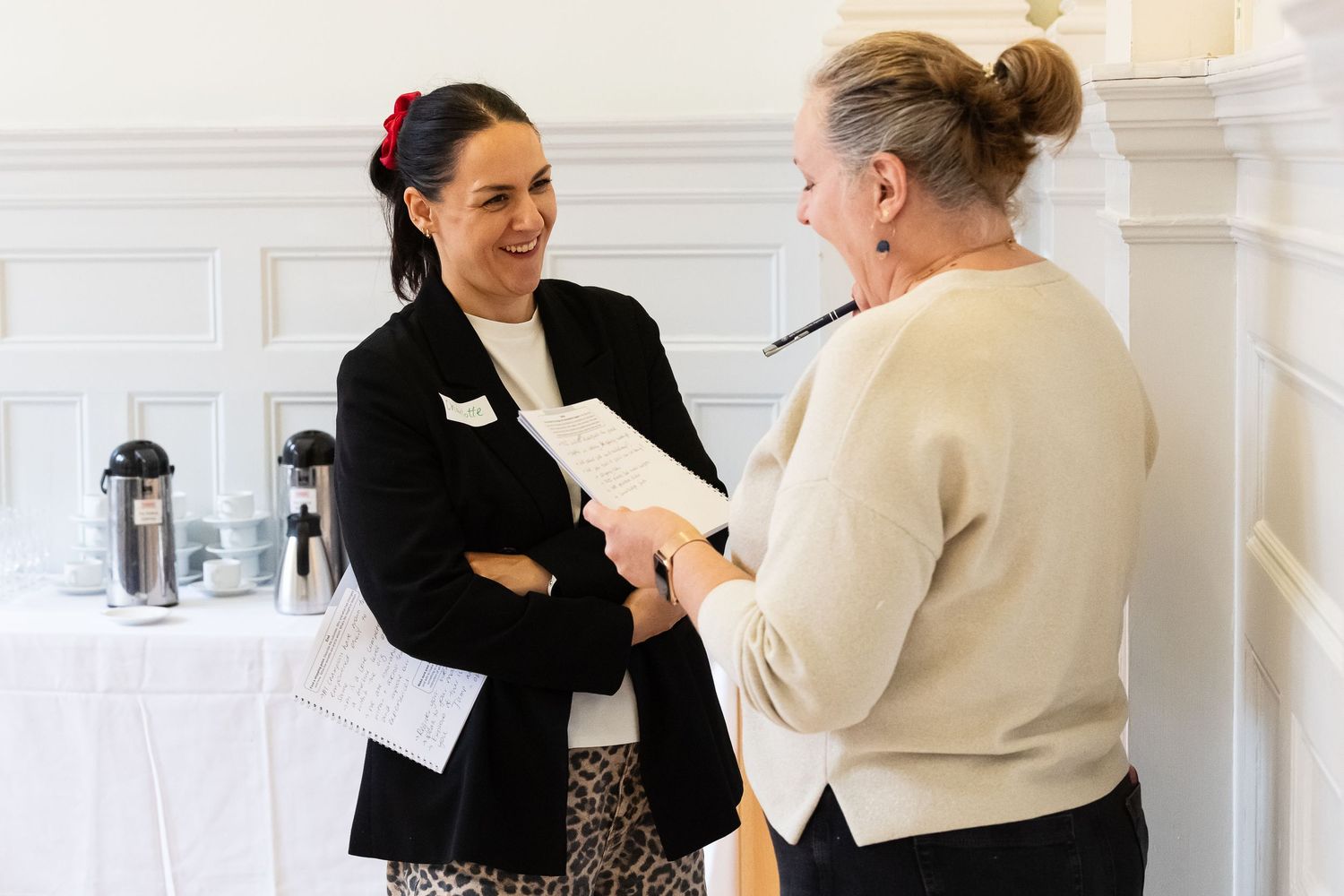 Two professional women smiling and exchanging documents at a formal indoor business event.