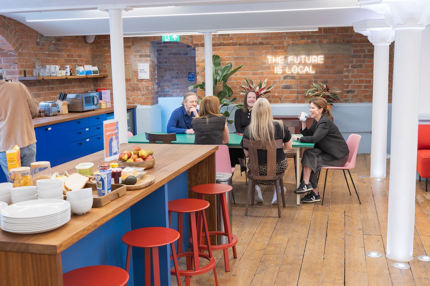 Modern co-working cafe space with red bar stools, brick wall neon sign reading The Future Is Local, and people collaborating.