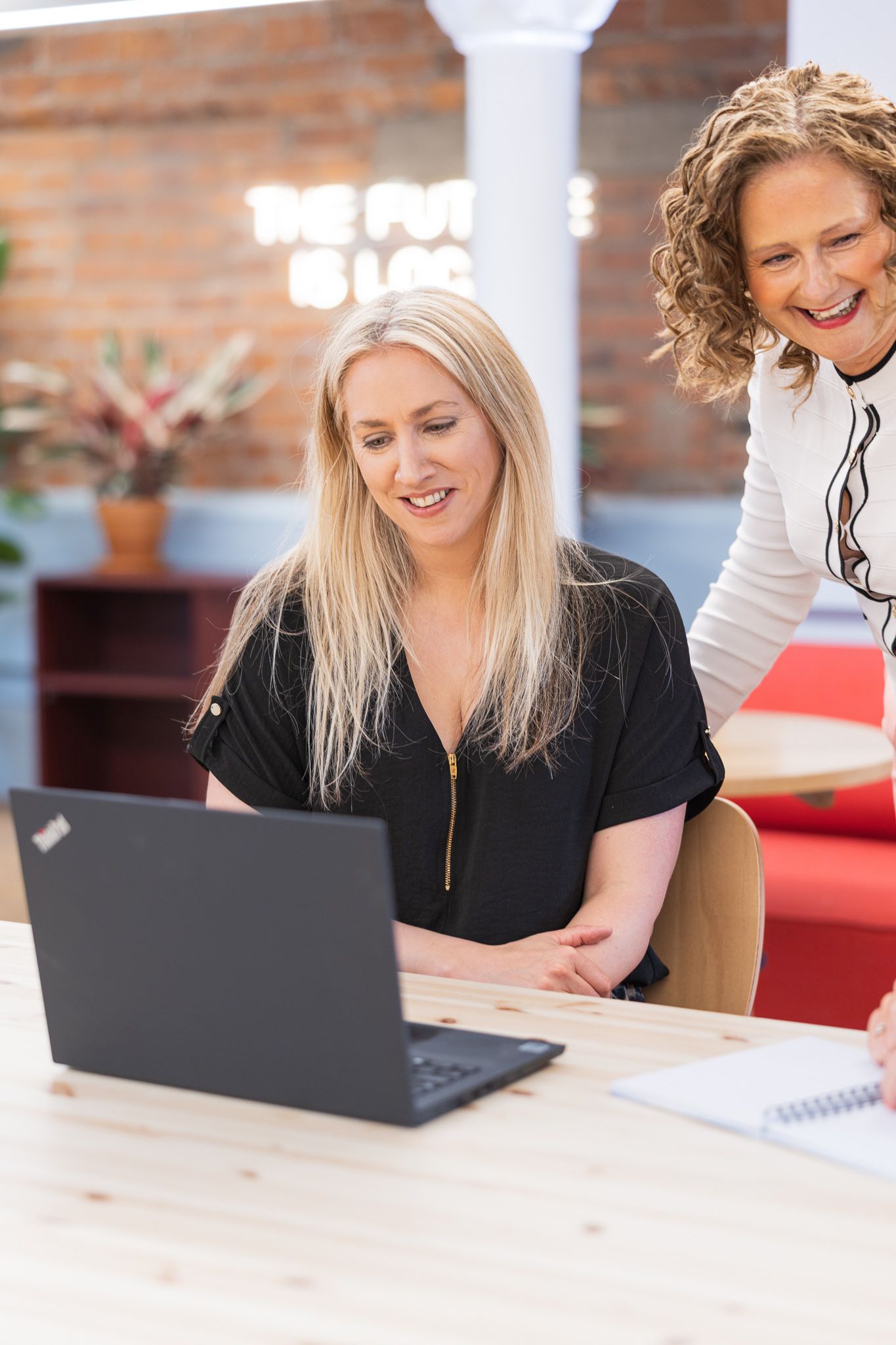 Two women smiling and collaborating on a Lenovo laptop at a wooden desk in a modern brick-walled office.