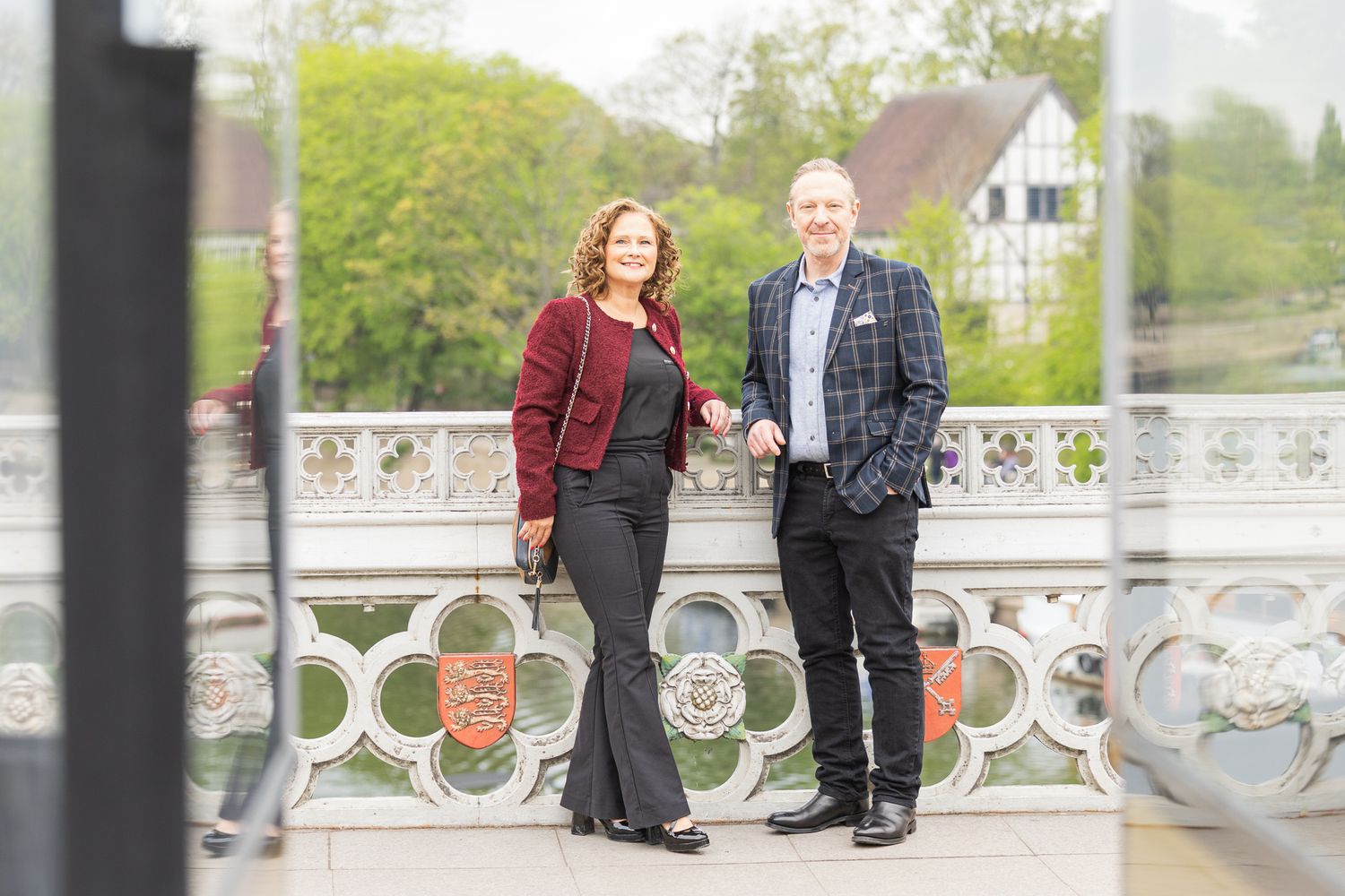 A man and woman pose together on an ornate bridge with colorful crests, surrounded by lush green trees.