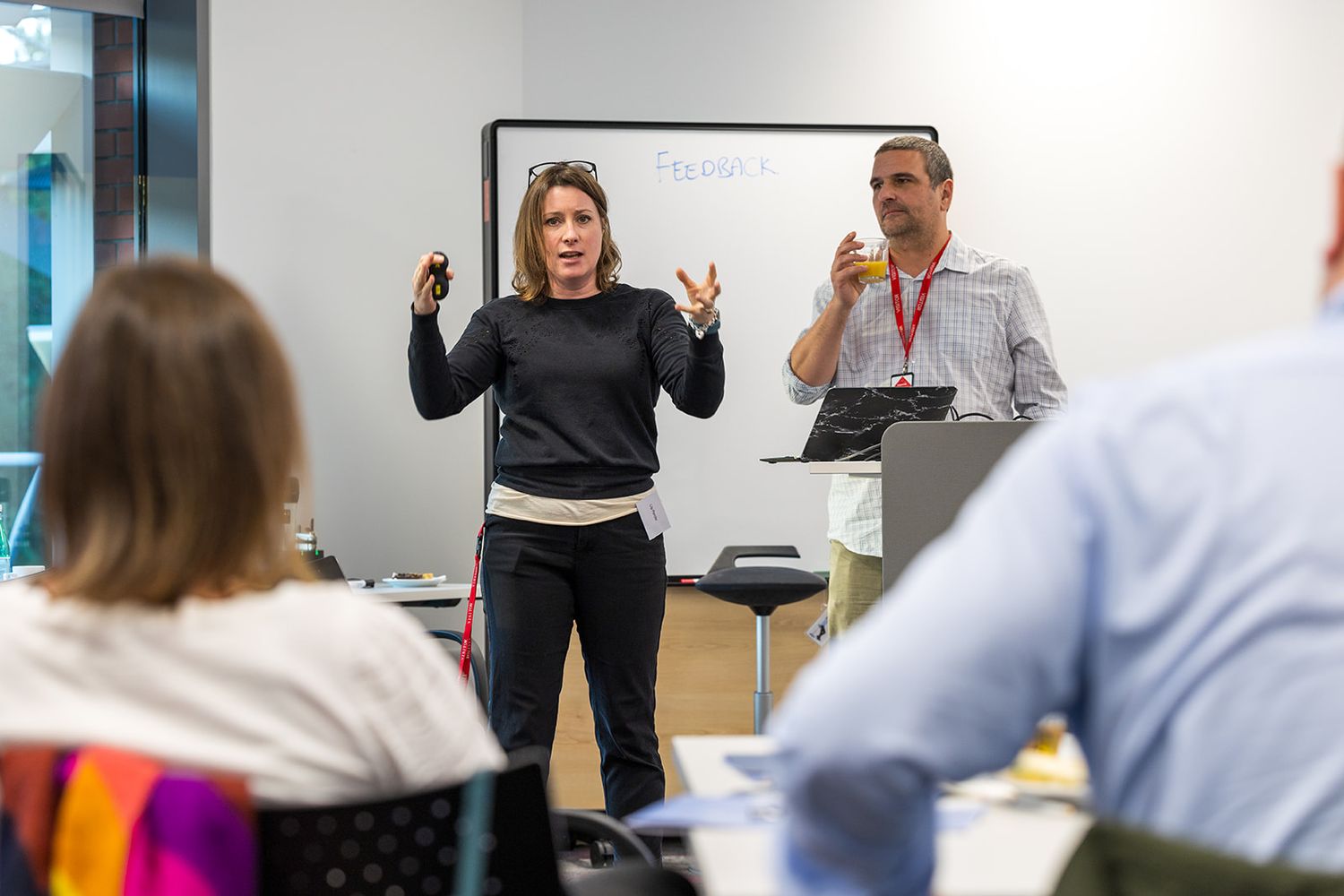 Two professionals presenting to colleagues in a modern office training room, with a whiteboard visible behind them.