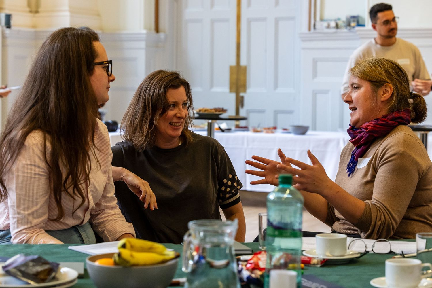 Three women engaged in lively conversation around a table with food and drinks in an elegant meeting room.