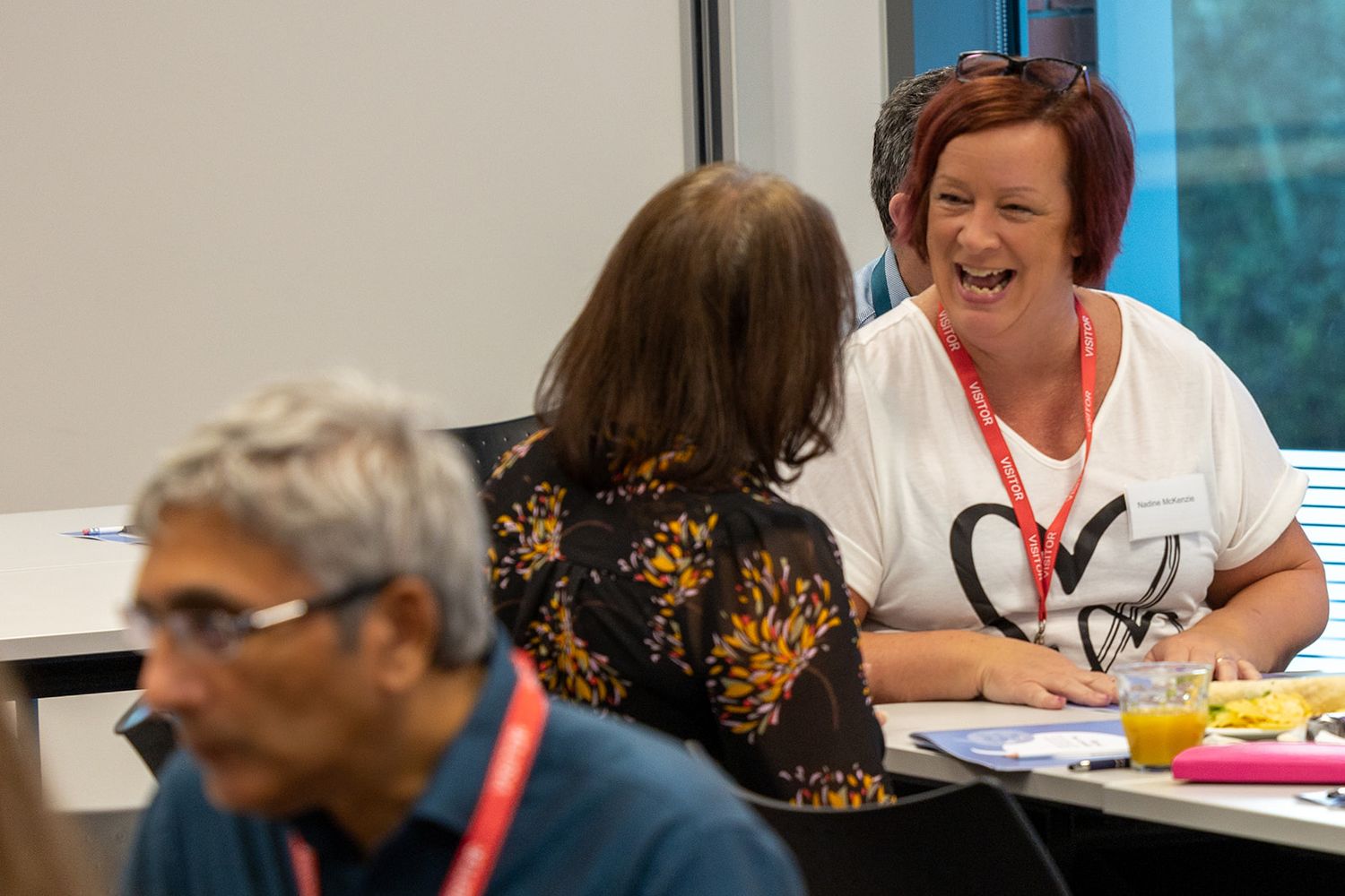 Woman laughing joyfully during a group meeting or workshop, wearing a red lanyard and white t-shirt.