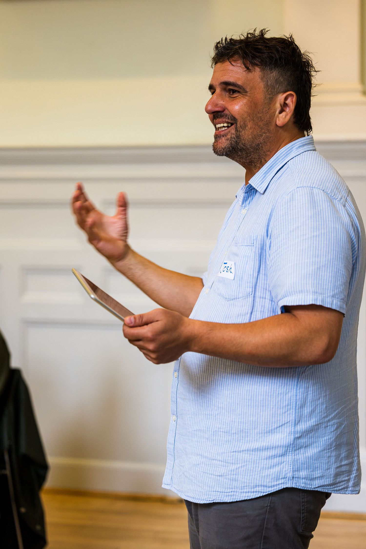 Smiling man in blue striped shirt holding tablet, gesturing while speaking at indoor event with name tag.