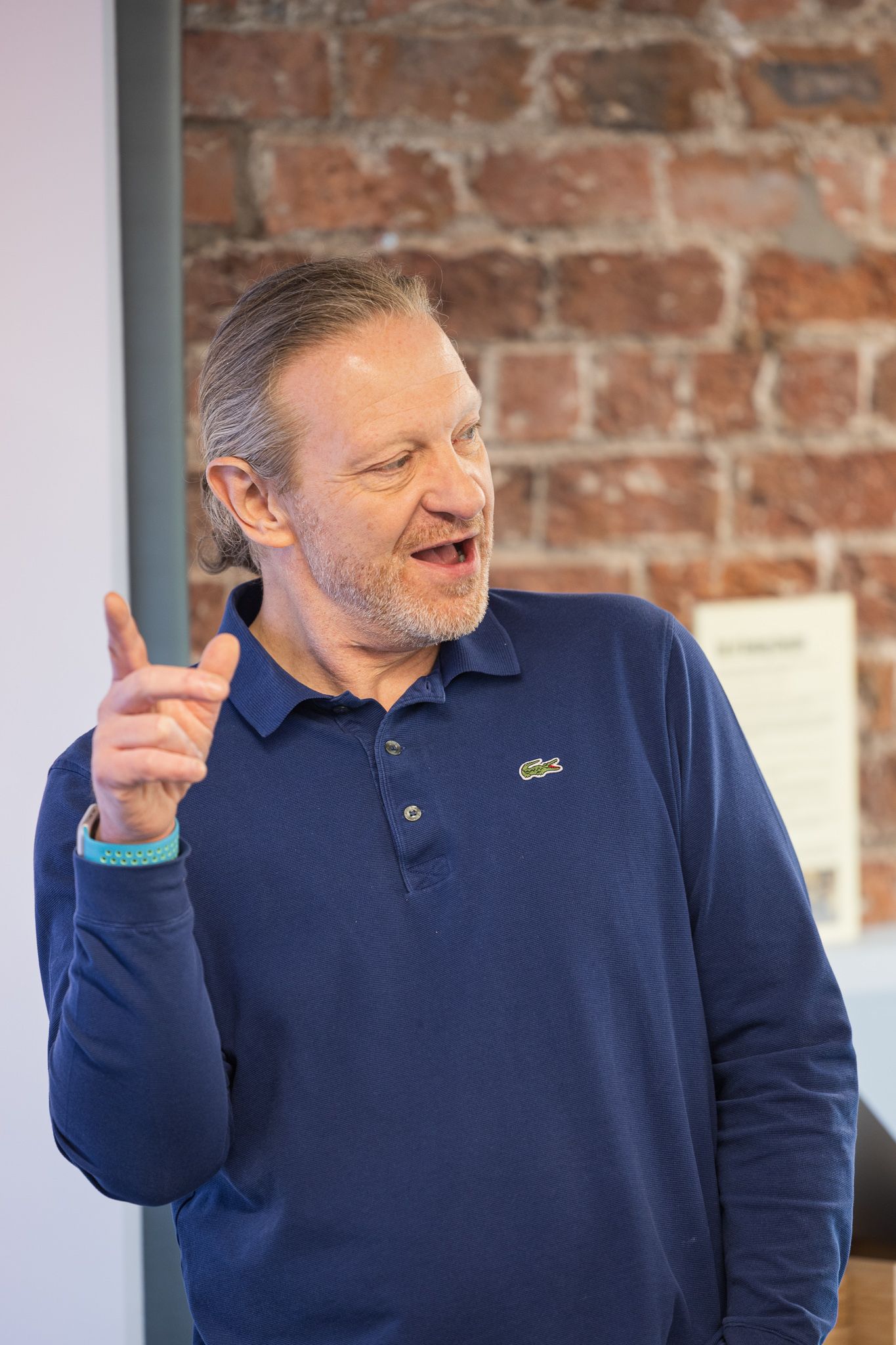 Middle-aged man in navy Lacoste polo shirt gesturing with finger raised, speaking expressively against brick wall backdrop.