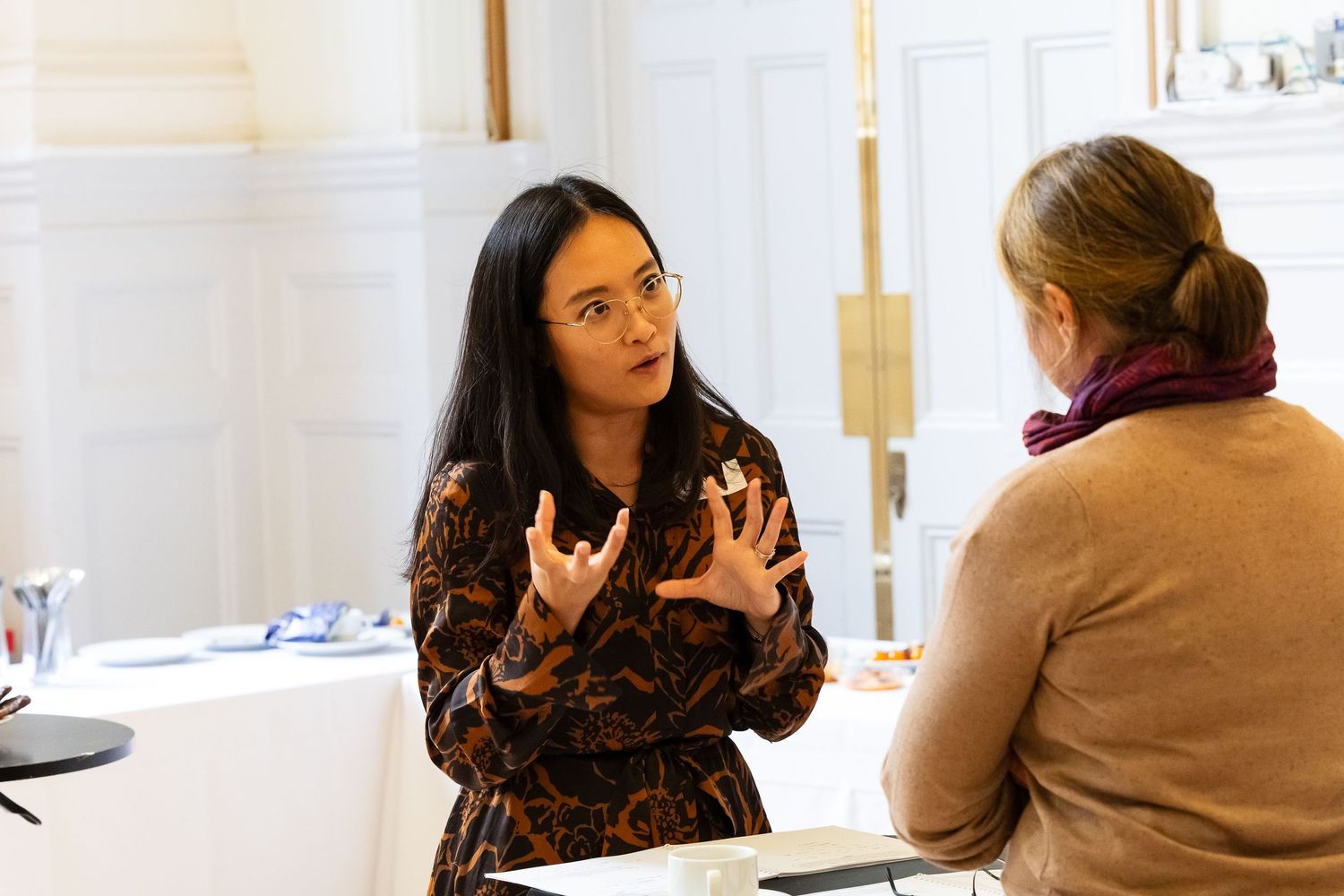 Two women engaged in conversation at an indoor event, one gesturing expressively while explaining something.